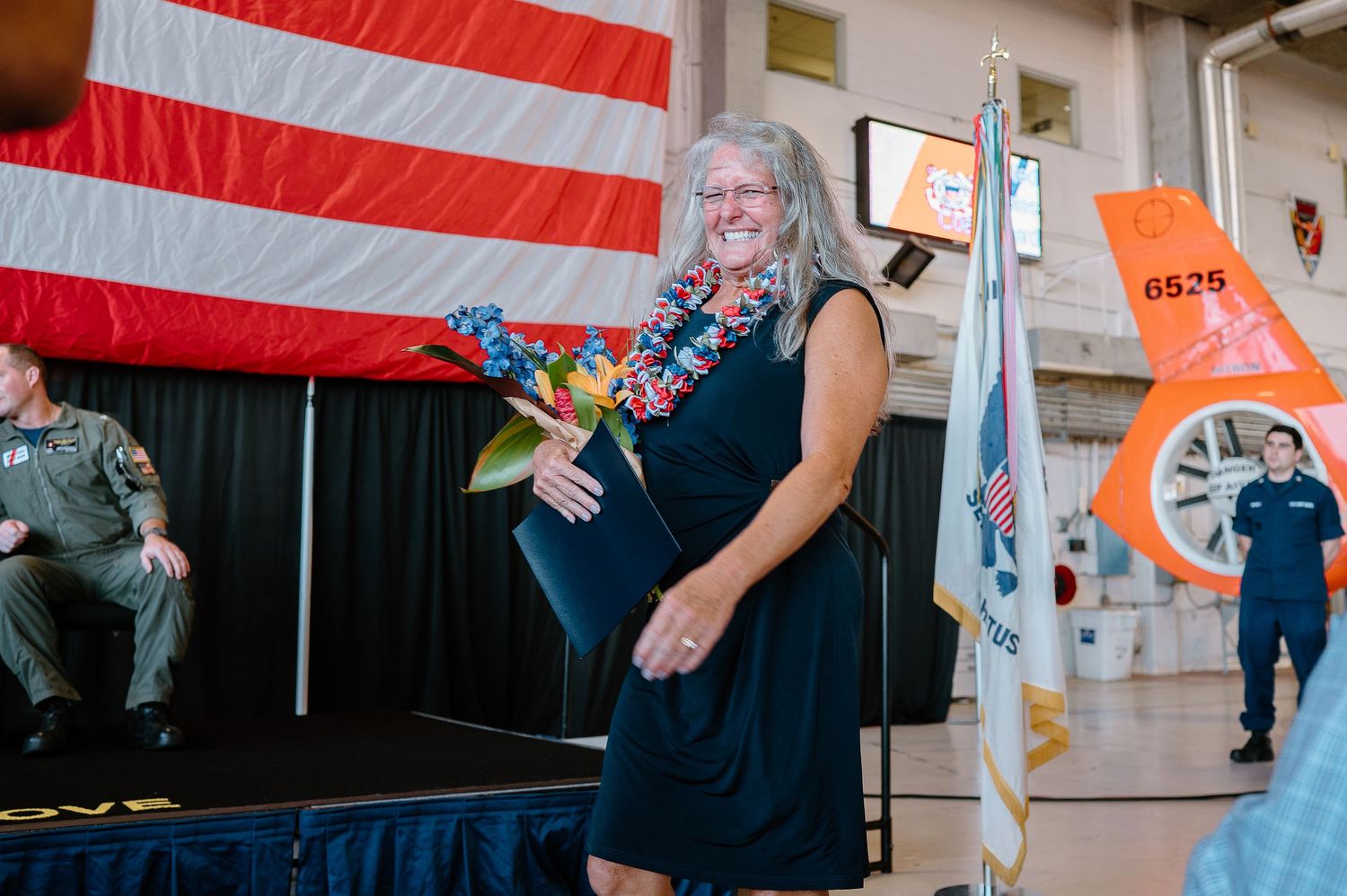 Woman in black dress holding flowers and lei smiles on stage at military ceremony with American flag backdrop.