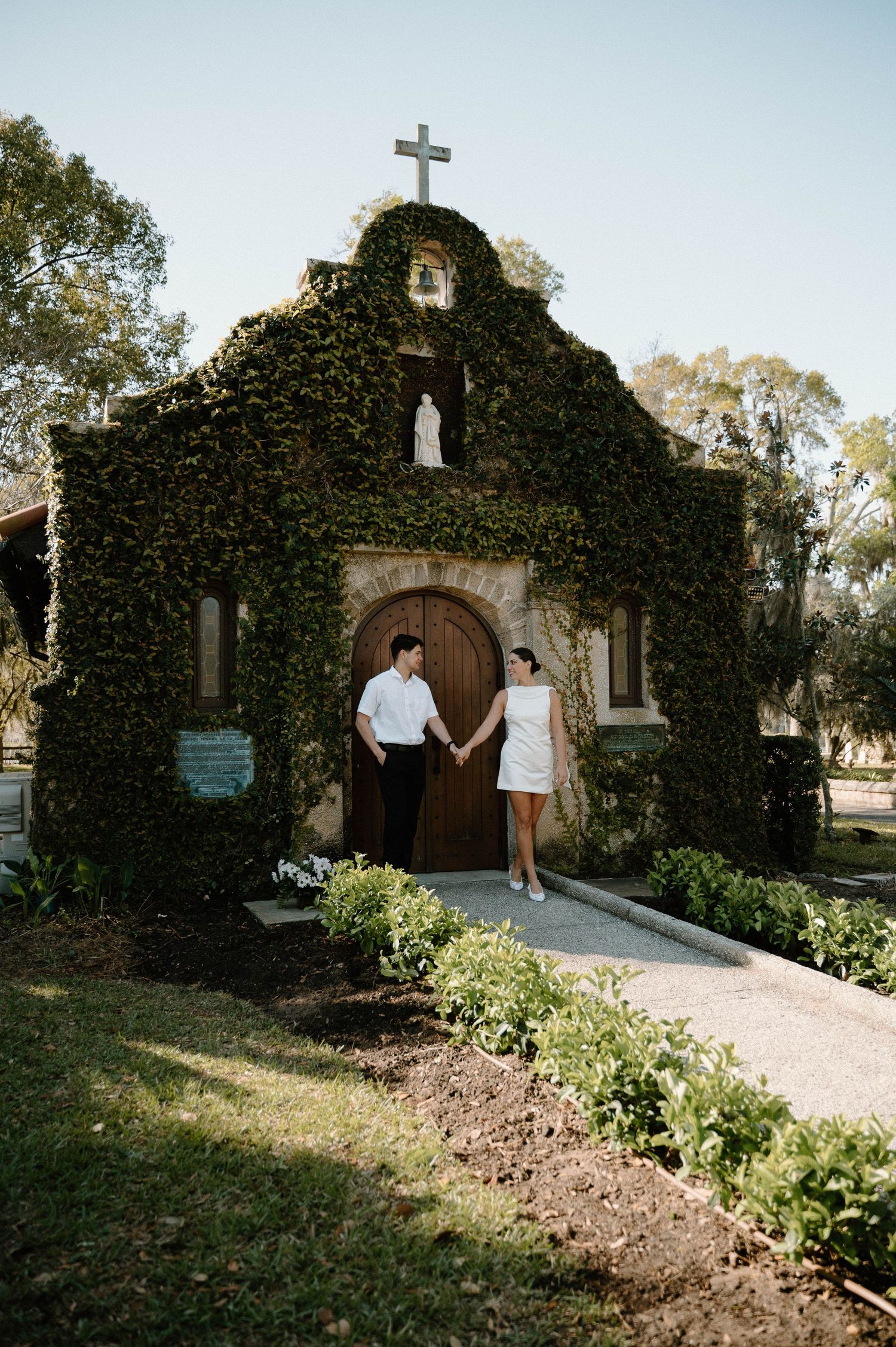 Couple holding hands outside an ivy-covered chapel with a cross, wearing white outfits on a sunny day.