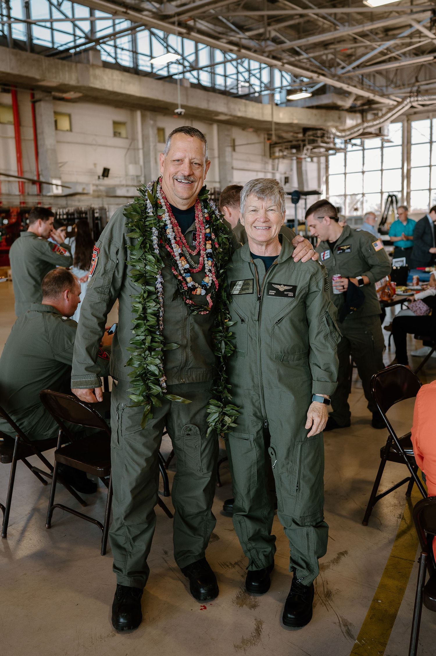 Two military personnel in olive flight suits smile together at an indoor aviation hangar event, one wearing Hawaiian leis.