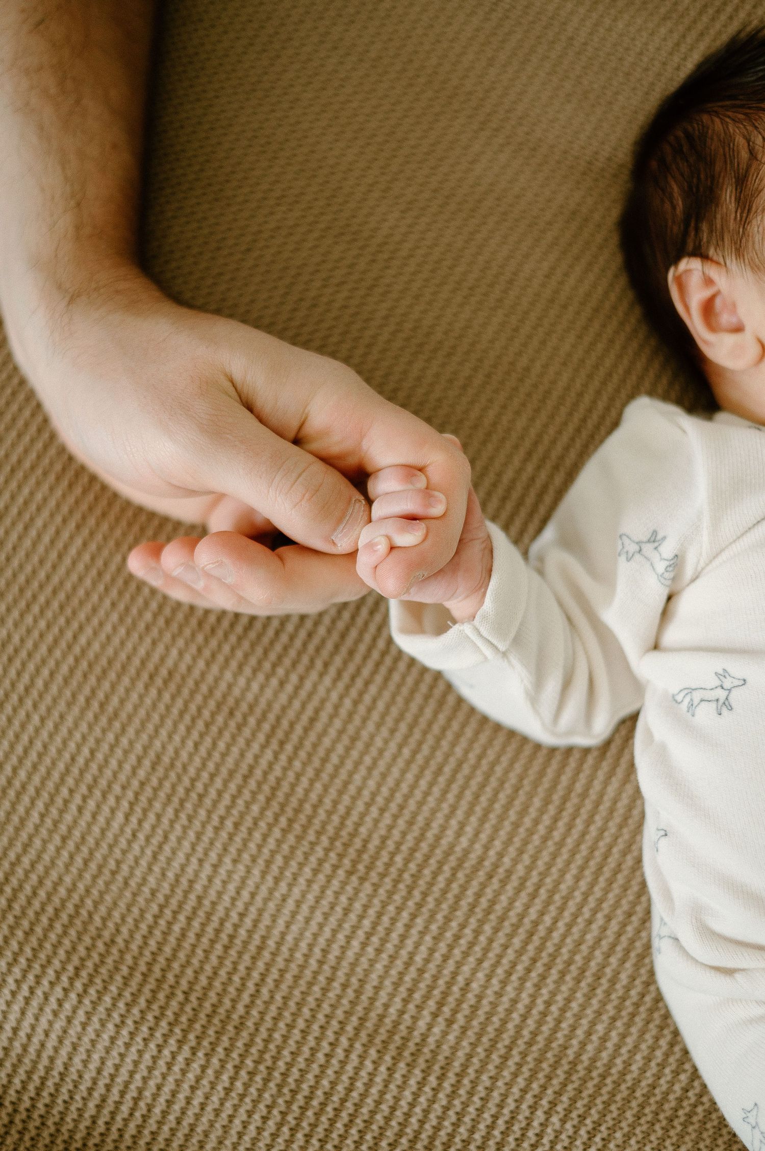 A tiny infant hand grasps an adult finger against a beige textured background.