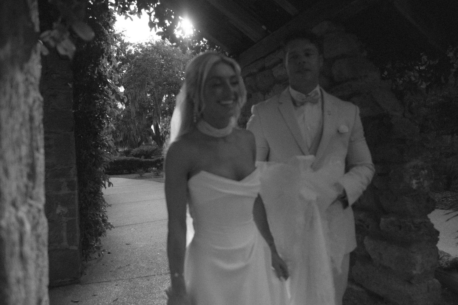 Black and white photo of a smiling bride in strapless gown and groom in white suit walking together outdoors.