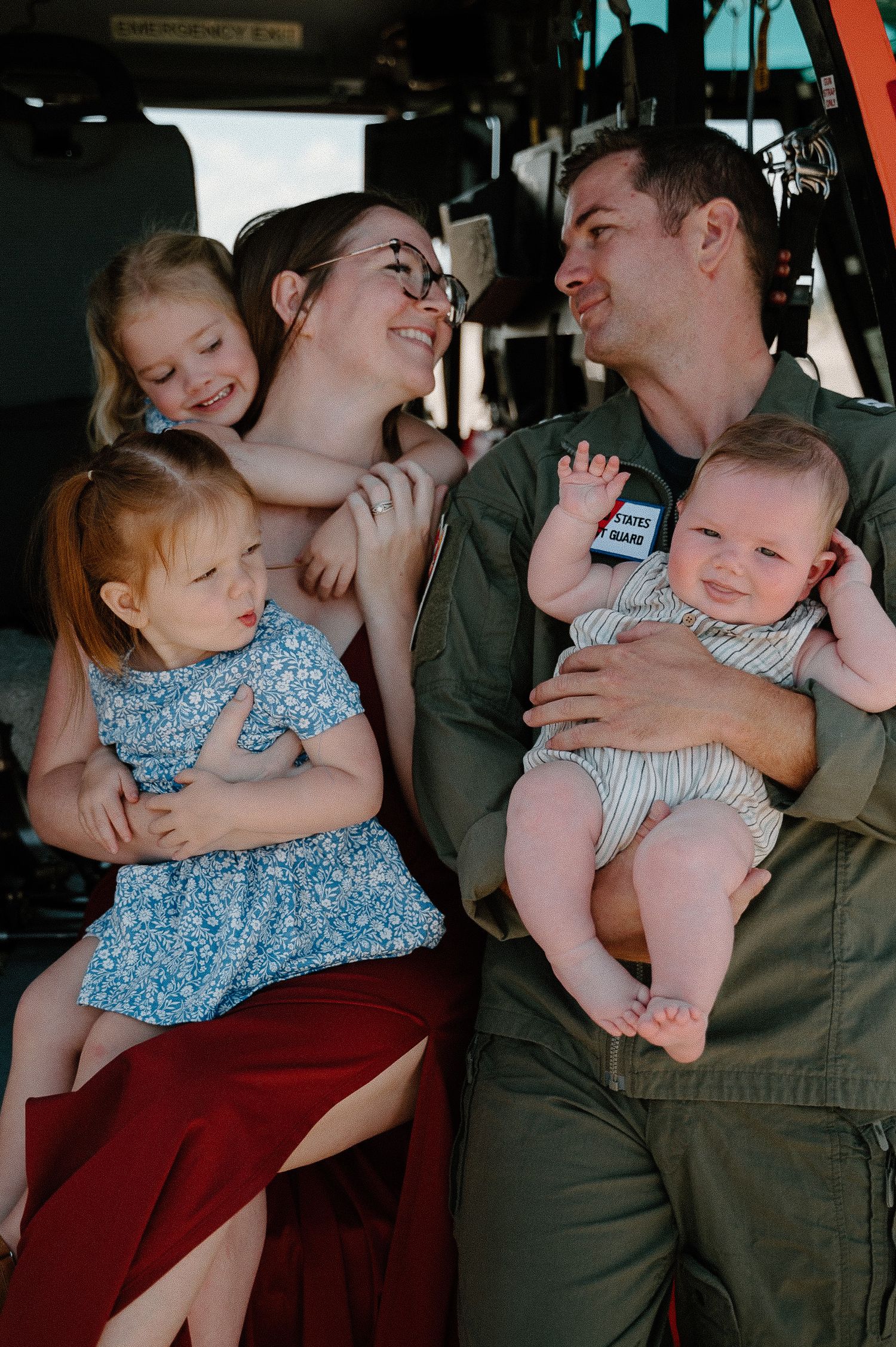 Military family with three young children smiling together inside a helicopter cockpit, father in green flight suit.