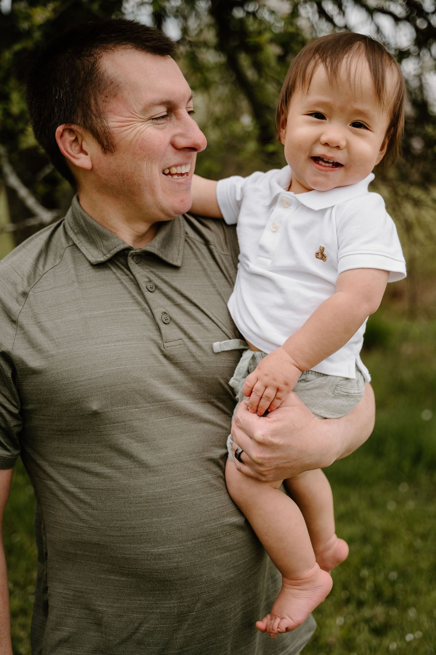 A smiling man in a green polo shirt holds a happy baby in a white onesie outdoors.