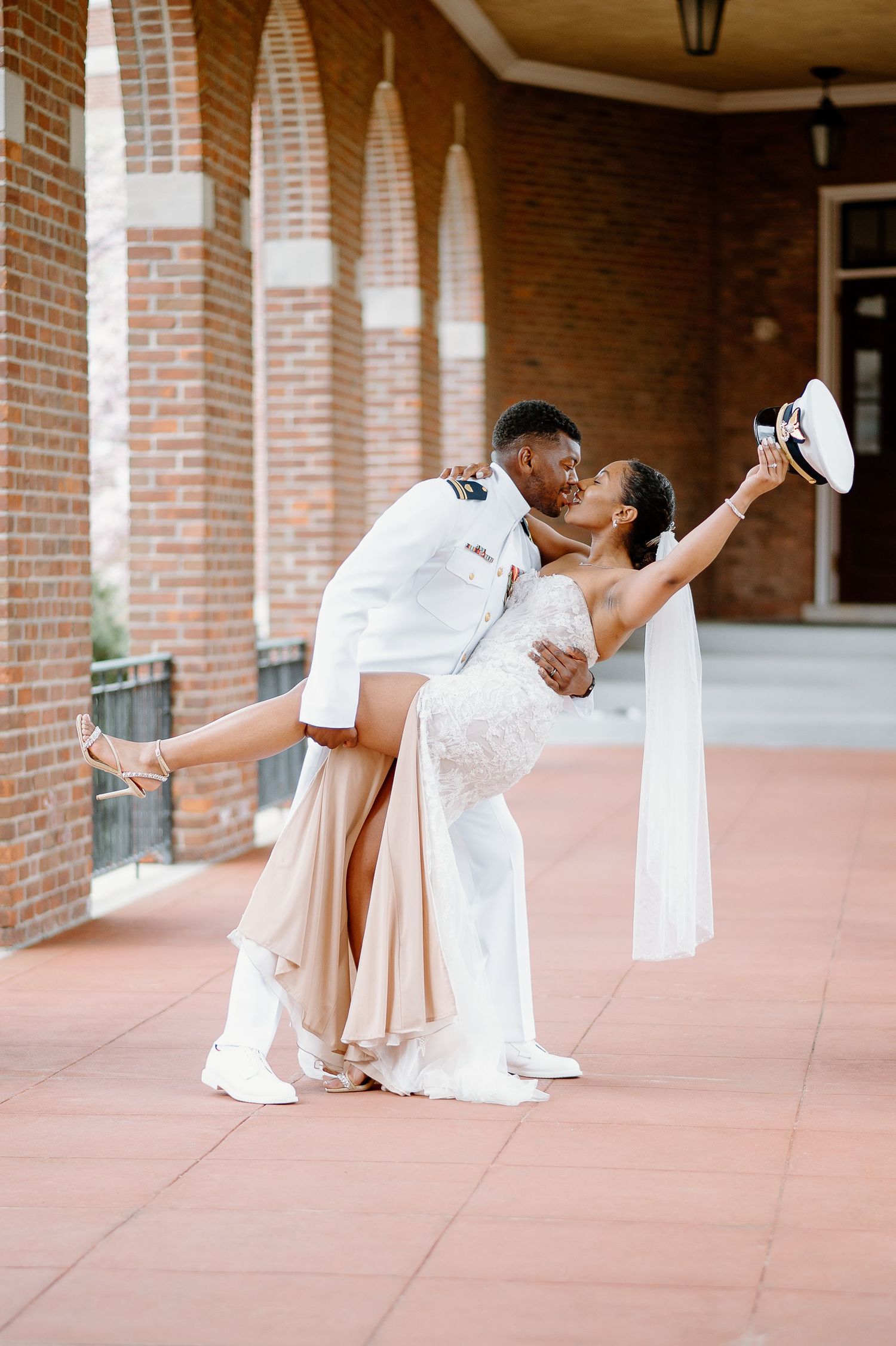 A couple in white wedding attire performs romantic dance poses under a brick colonnade.