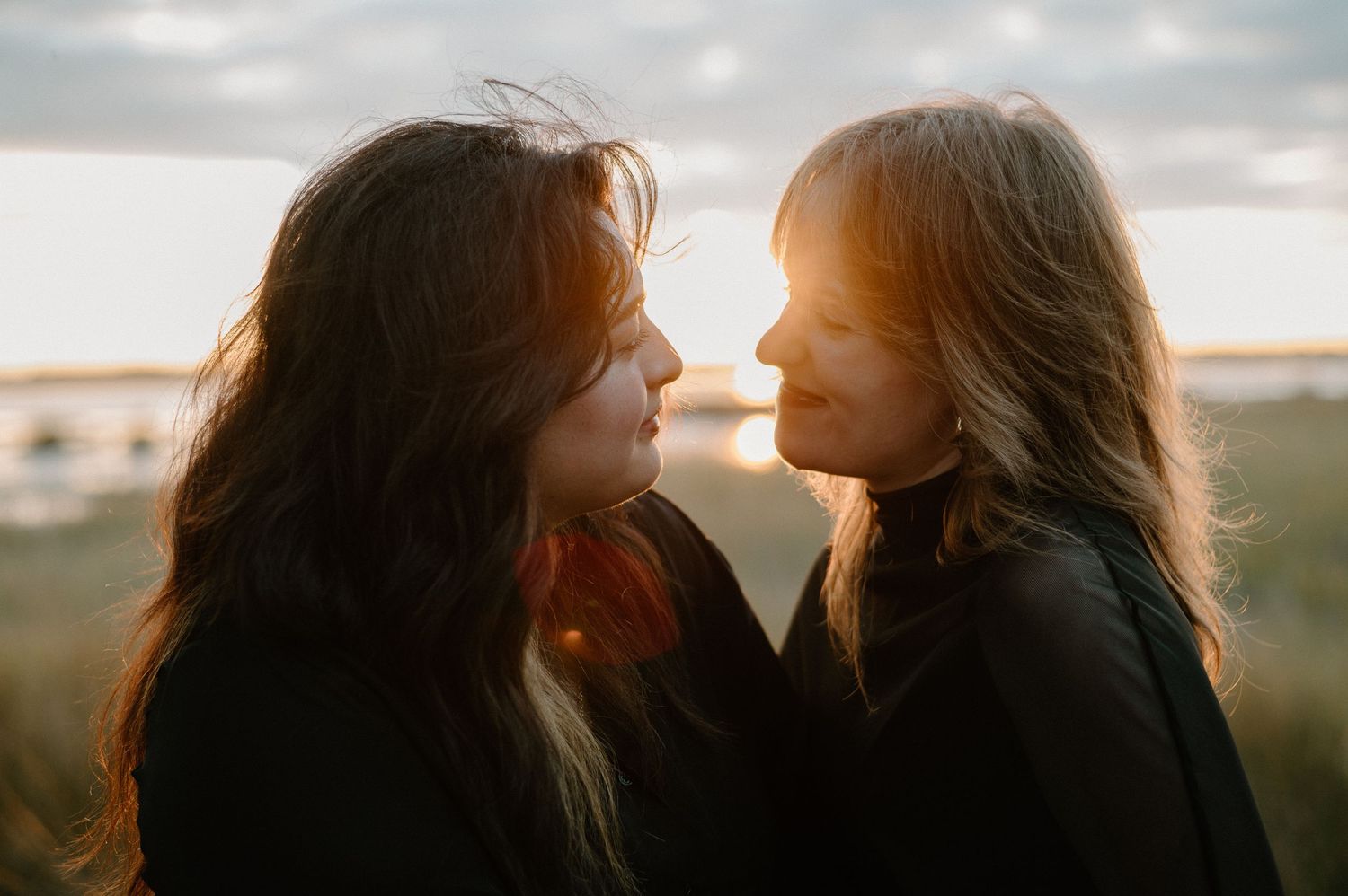 Two women with long dark hair face each other at golden hour outdoors, bathed in warm sunset light. Jekyll island engagement photographer