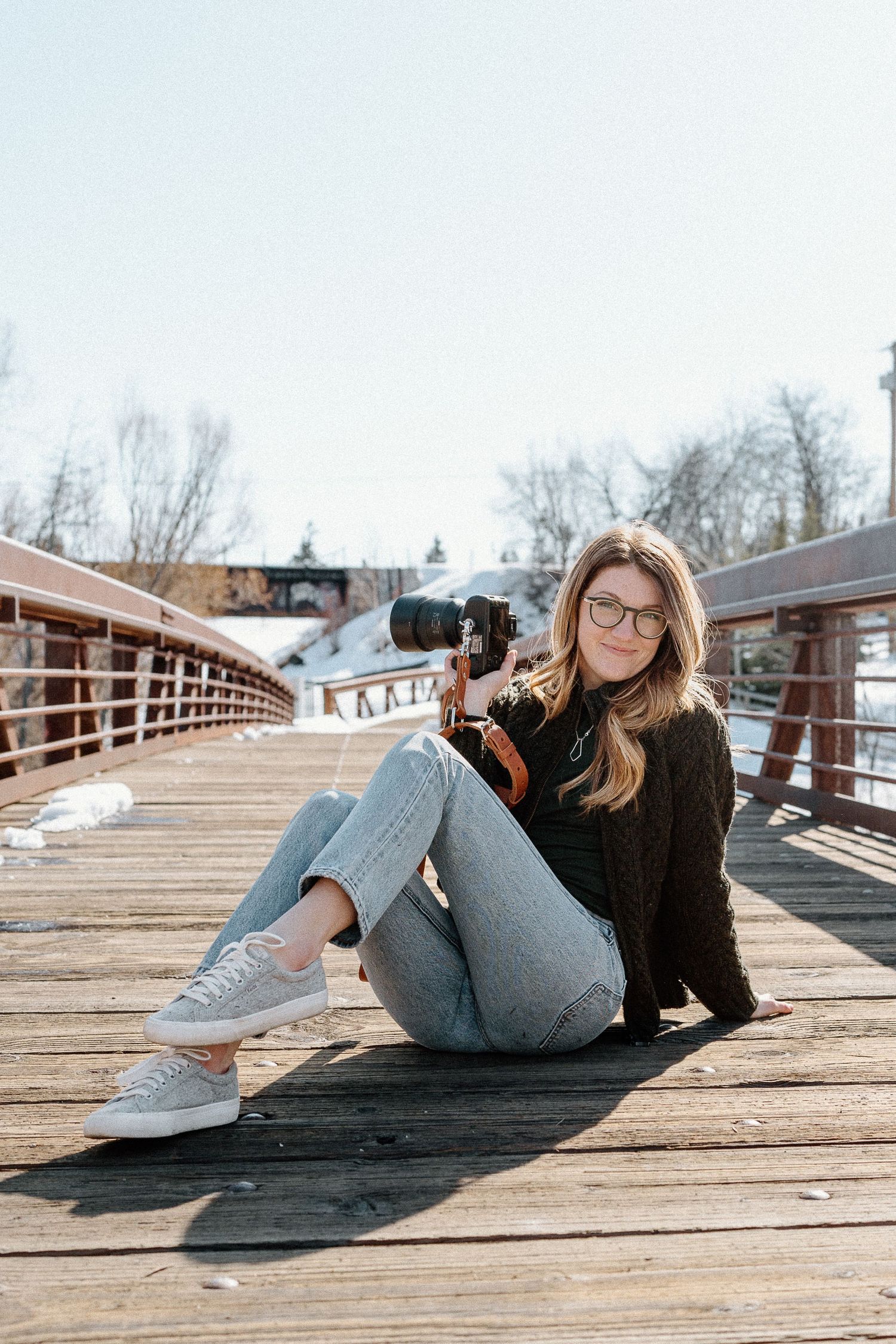 A person in casual attire lies on a wooden bridge deck while taking photos with a camera on a cloudy day.