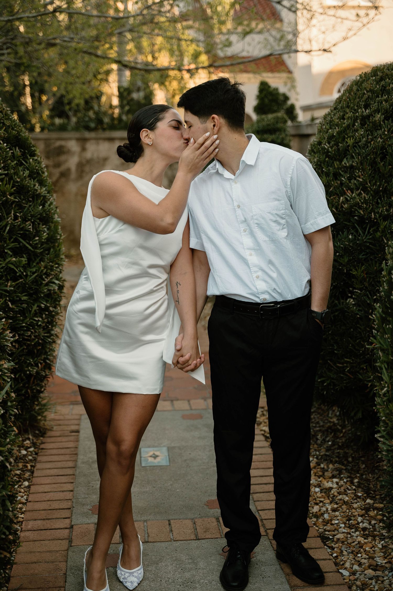 Couple sharing a romantic kiss in a garden pathway, woman in white mini dress holding hands with man in white shirt.