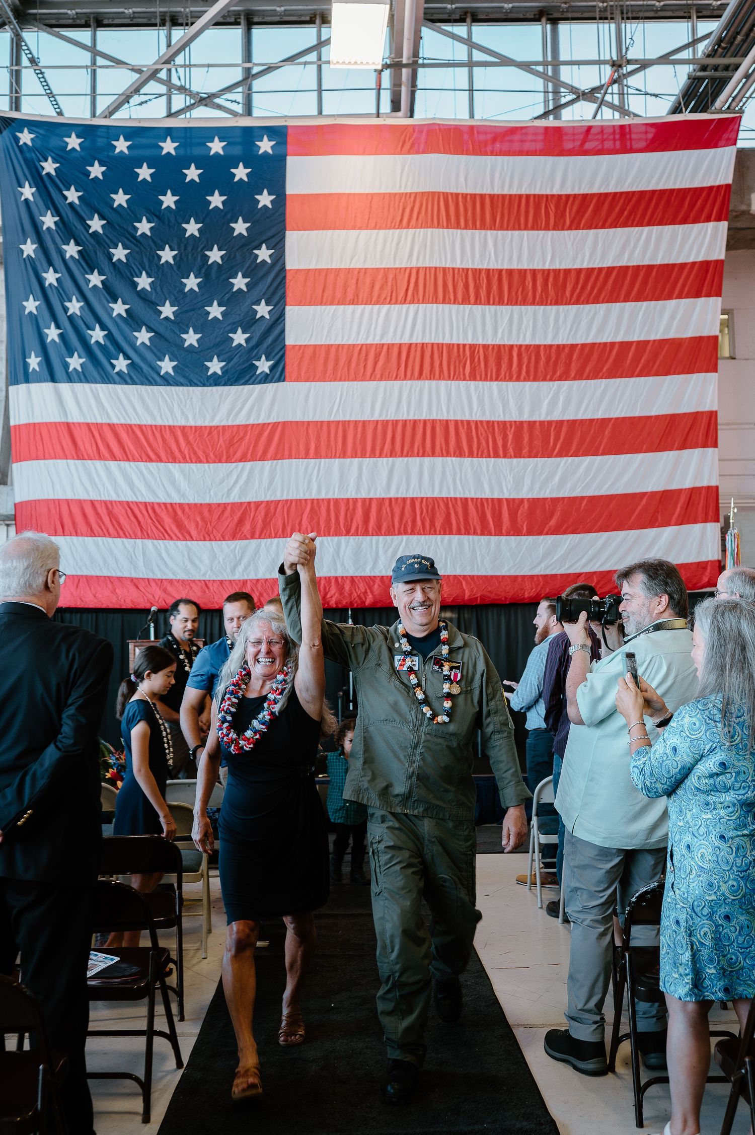 Veteran in flight suit raises hand with woman before large American flag at military retirement ceremony.