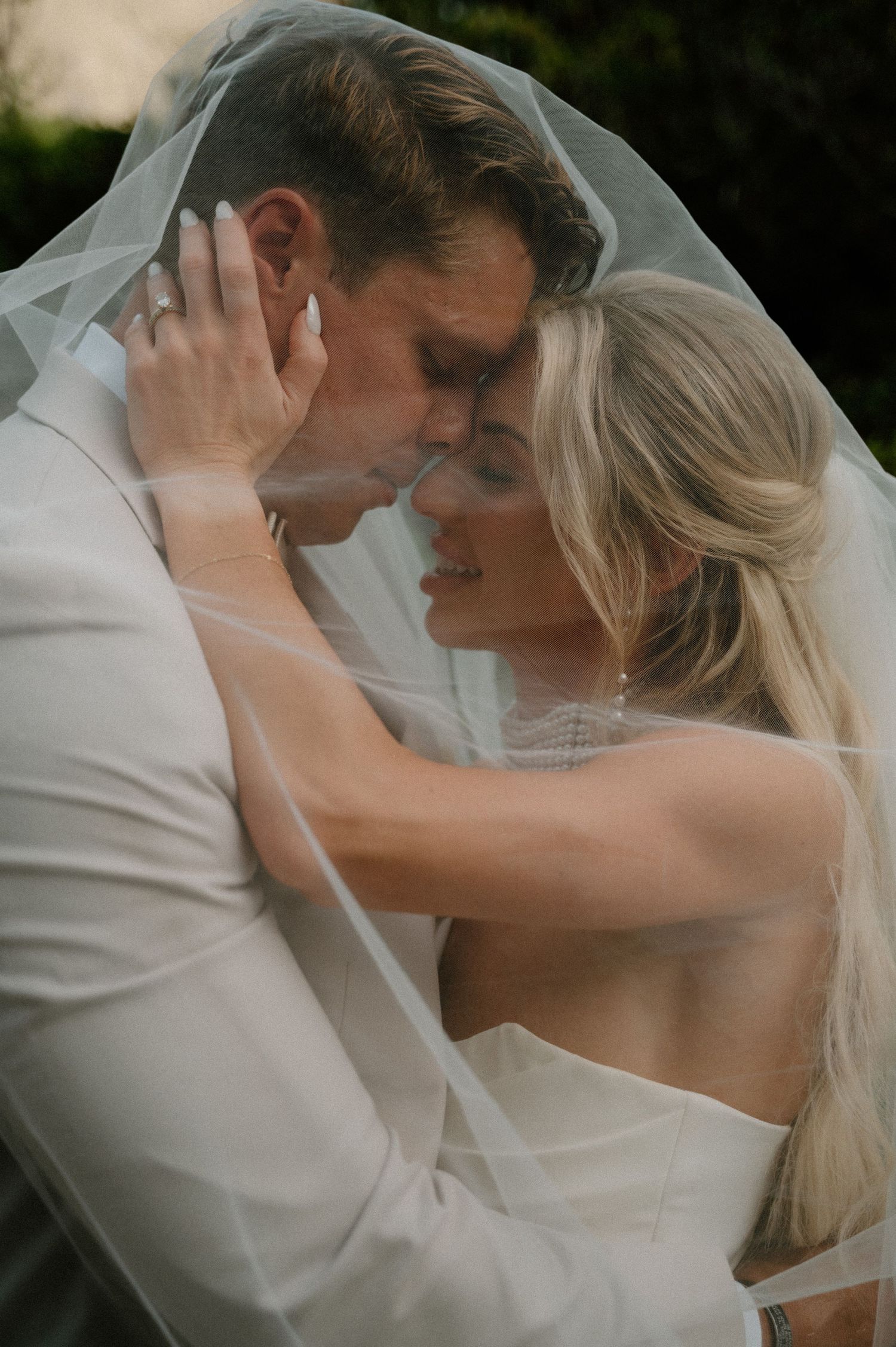 A bride and groom share an intimate moment under a flowing wedding veil, their foreheads touching tenderly.