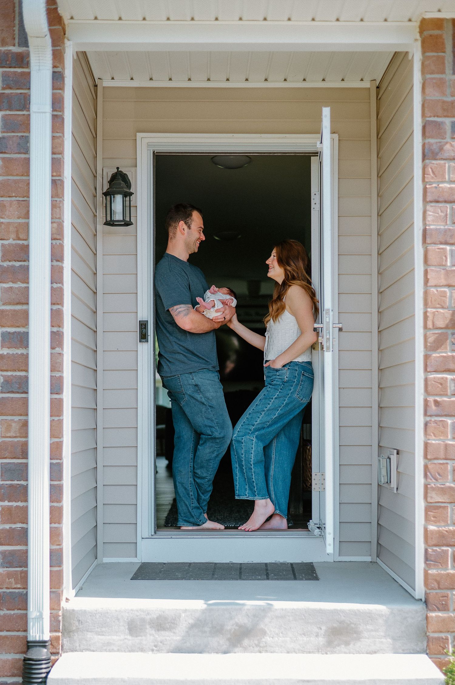 A couple stands together in a doorway entrance of a home with tan siding and brick accents.