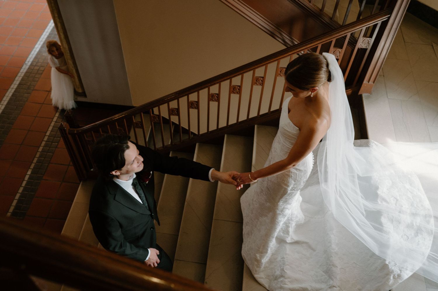 Bride in white gown holds hands with groom on elegant staircase, captured from above in a romantic wedding moment. The masonic downtown DTW