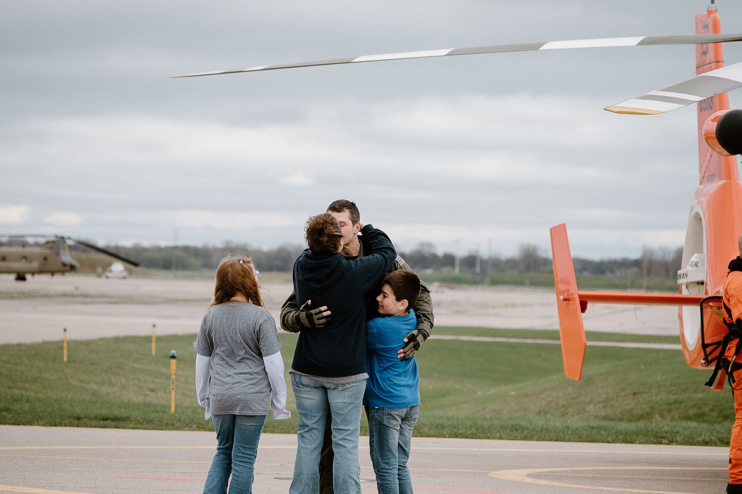 A small group embraces while standing near an orange helicopter on the tarmac.