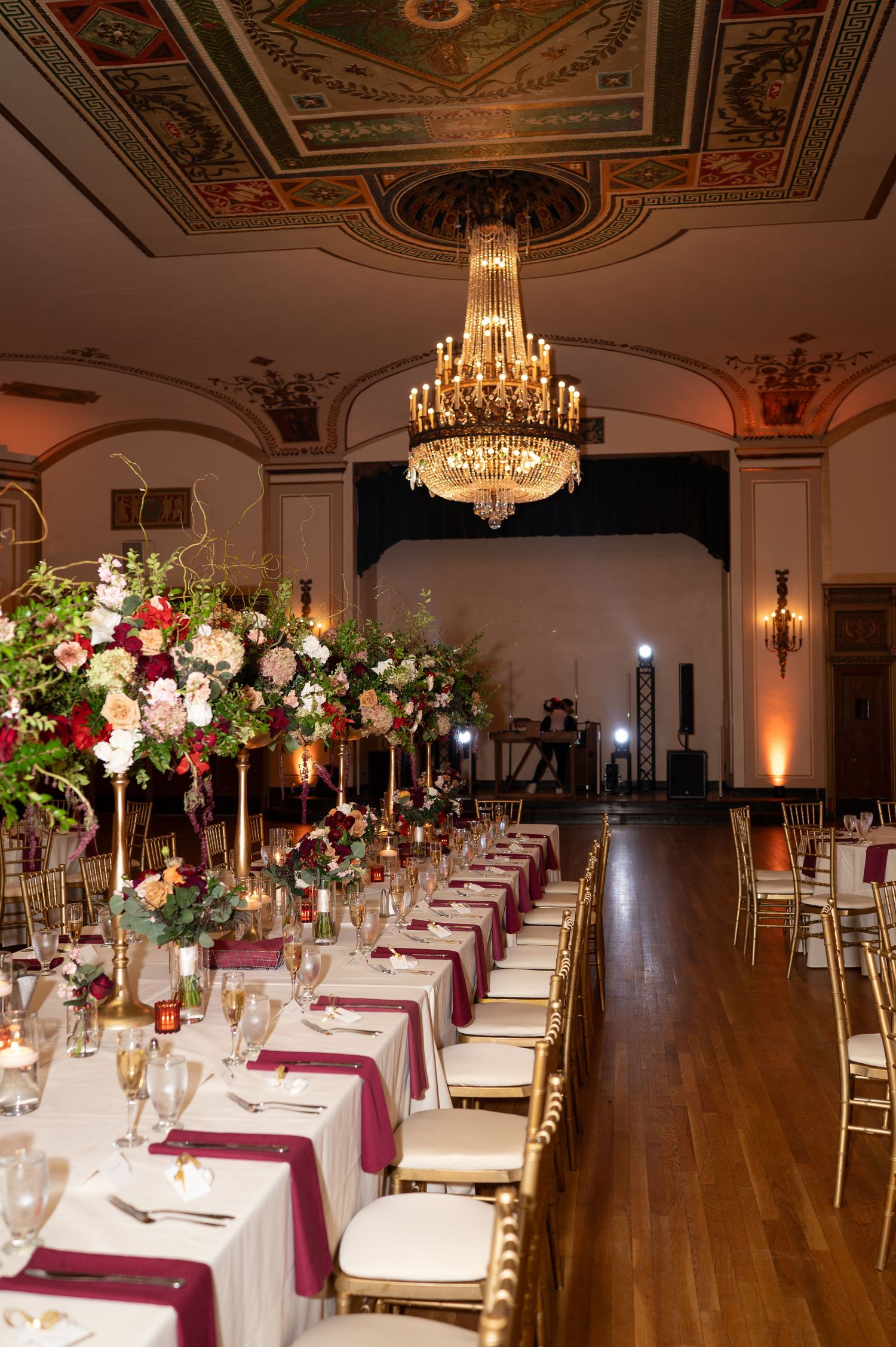 Elegant wedding reception venue with long tables, burgundy linens, crystal chandeliers, and tall floral centerpieces.