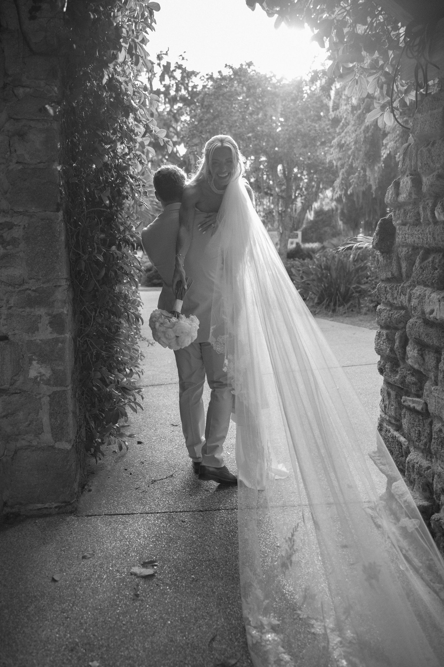 Black and white photo of a bride in a long veil being lifted by groom near stone wall outdoors.