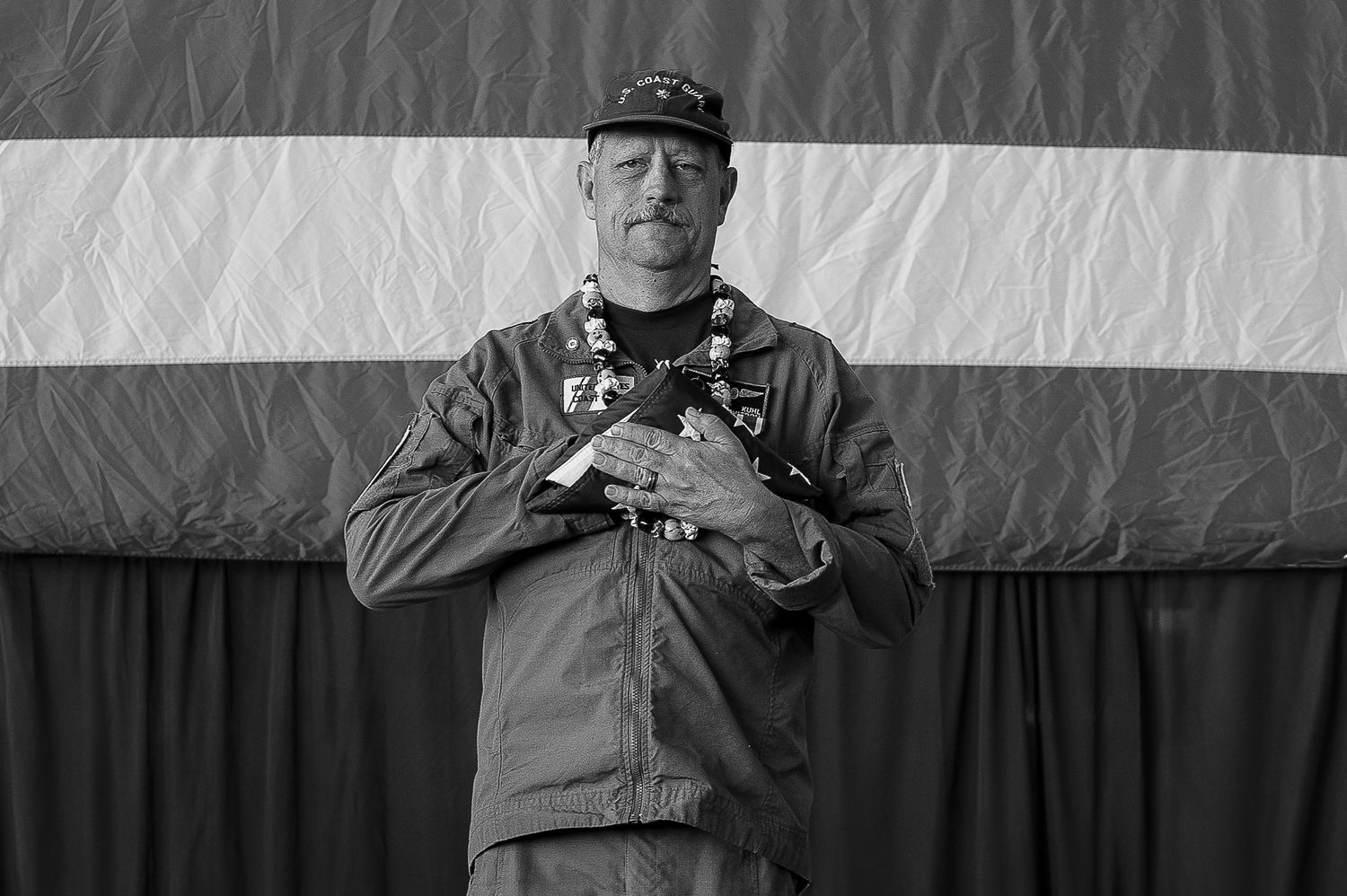 Black and white photo of a man in military uniform with lei, standing before a striped flag with hands crossed.
