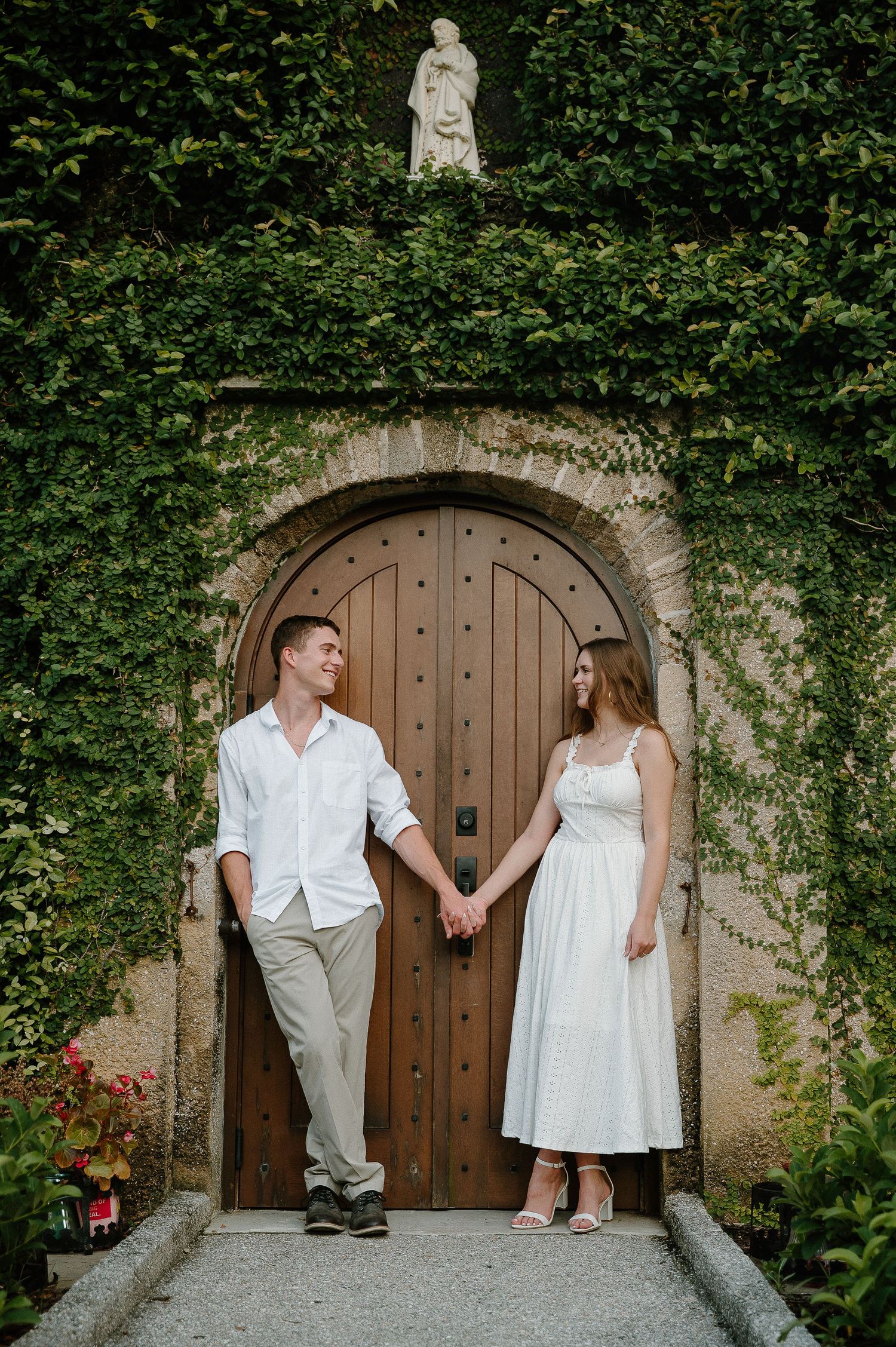 Couple holding hands in white outfits standing before an ivy-covered arched wooden door with a stone statue above.