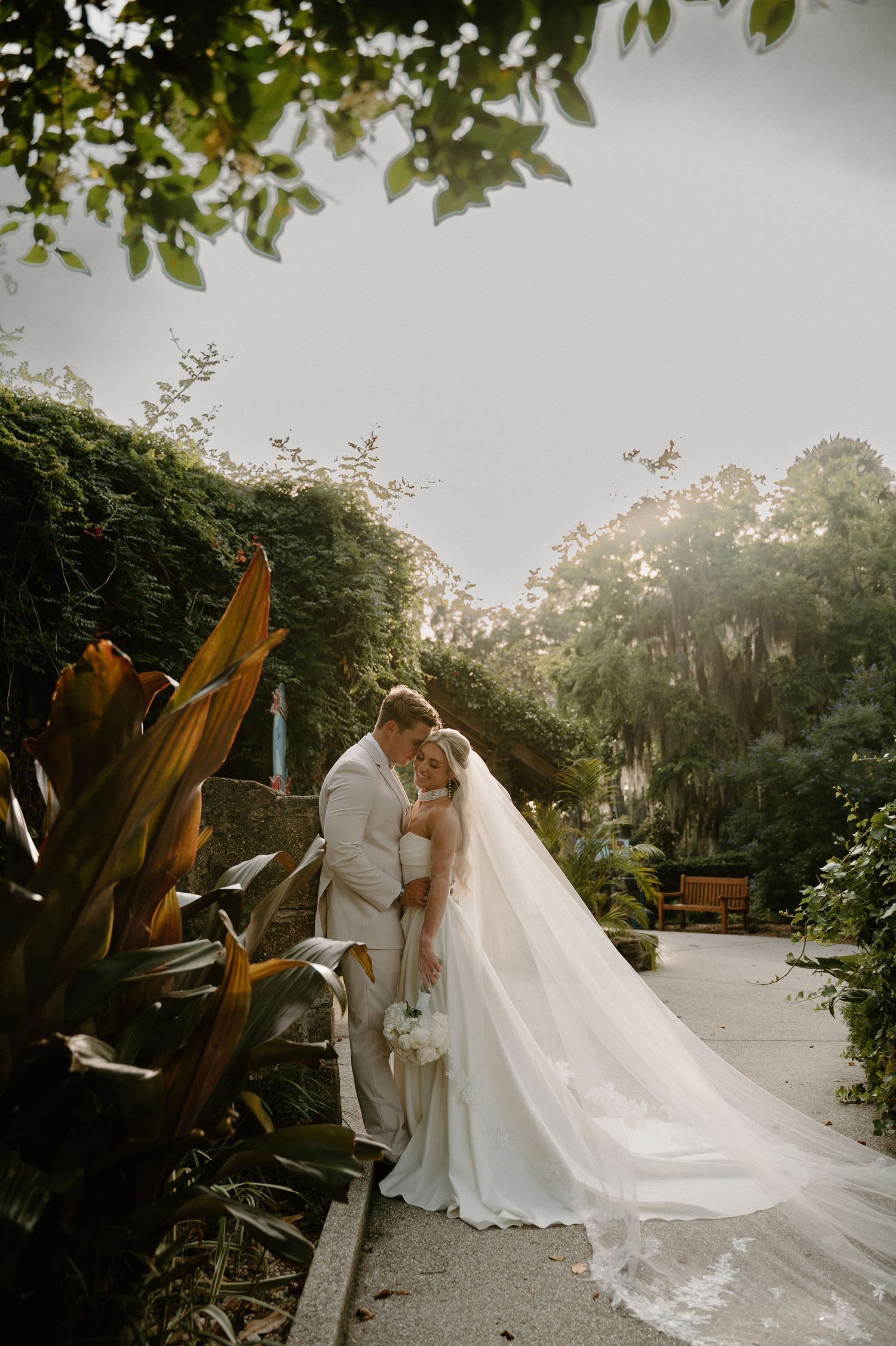 Bride and groom share a romantic embrace on a garden path, her cathedral veil flowing gracefully amid lush greenery.
