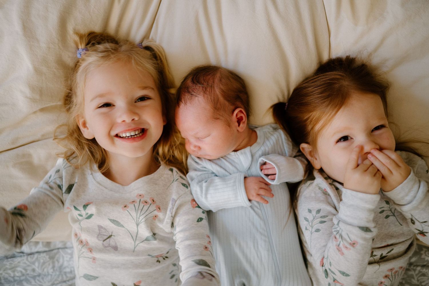 Three young siblings, including a newborn baby, lying together on a white bed, smiling and cuddling in matching pajamas.
