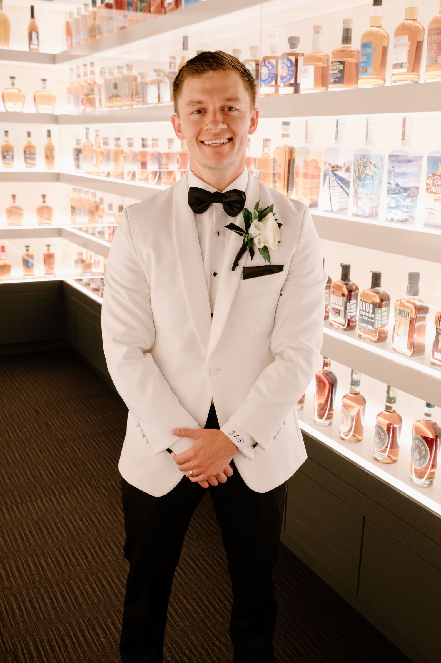 Person in white tuxedo jacket posing against illuminated liquor bottle display shelves.