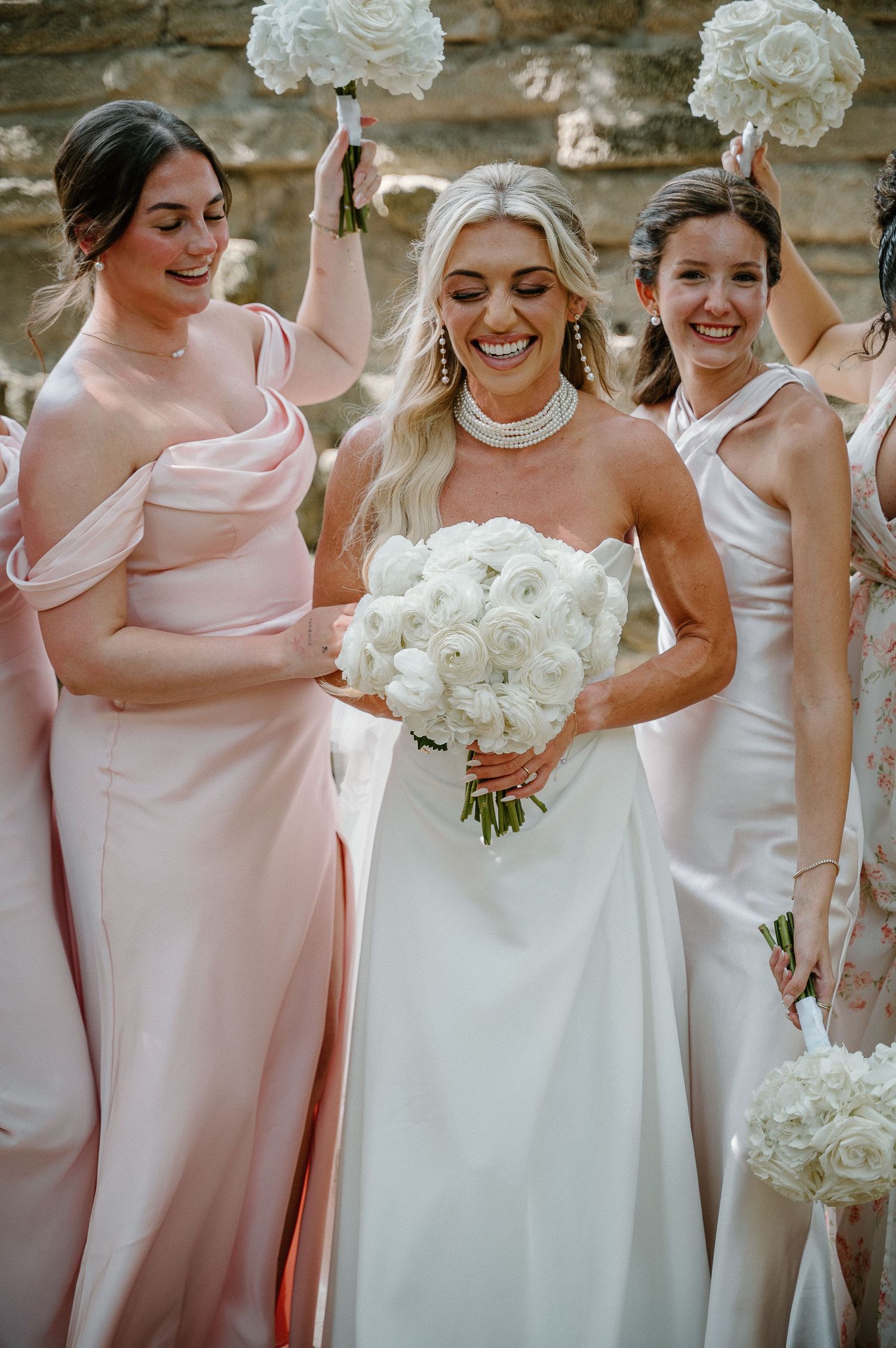 Bride in white strapless gown holding white ranunculus bouquet, flanked by bridesmaids in blush pink satin dresses.
