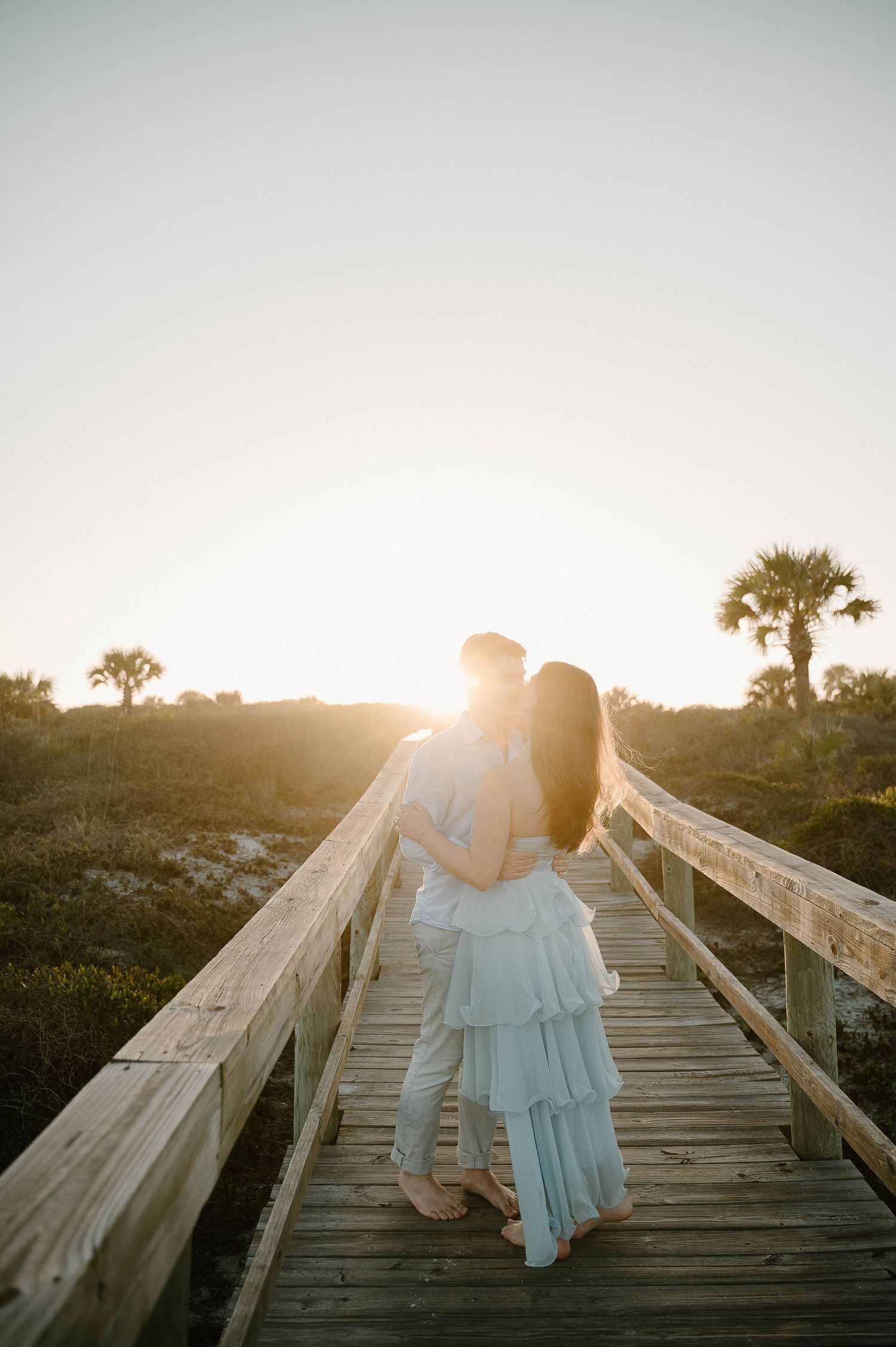Couple embracing on a wooden boardwalk at golden sunset with palm trees, wearing light blue and white outfits.