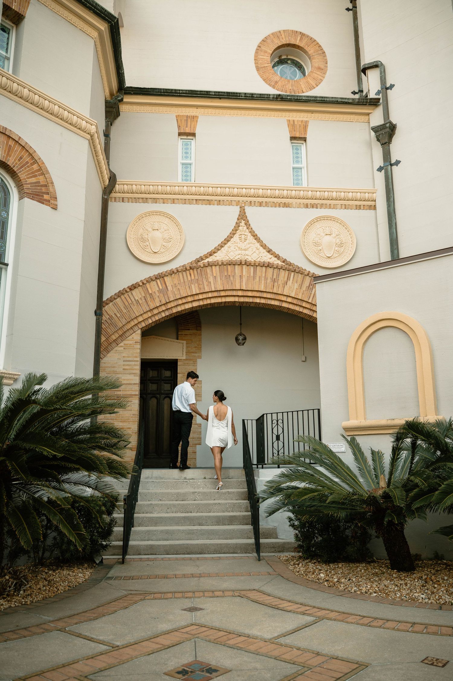 Couple in wedding attire holding hands walking up stairs toward elegant building entrance with arched brick doorway and palm trees.