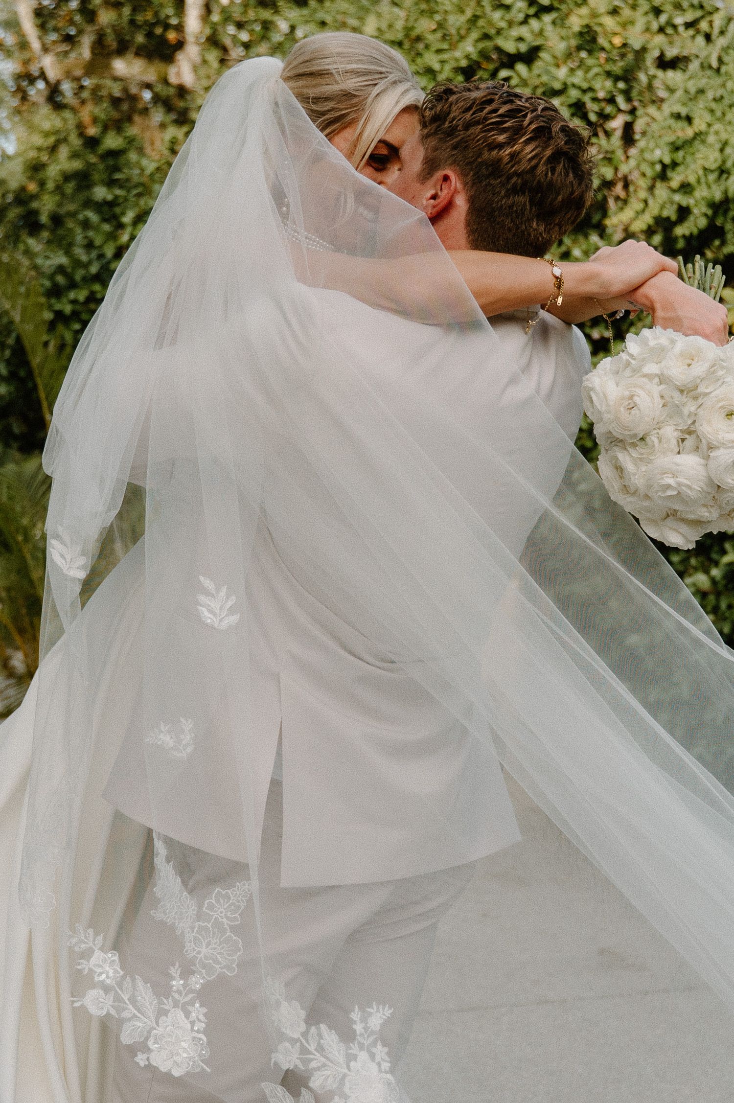 Bride and groom share a romantic kiss, her floral lace veil cascading around them as she holds a white bouquet.