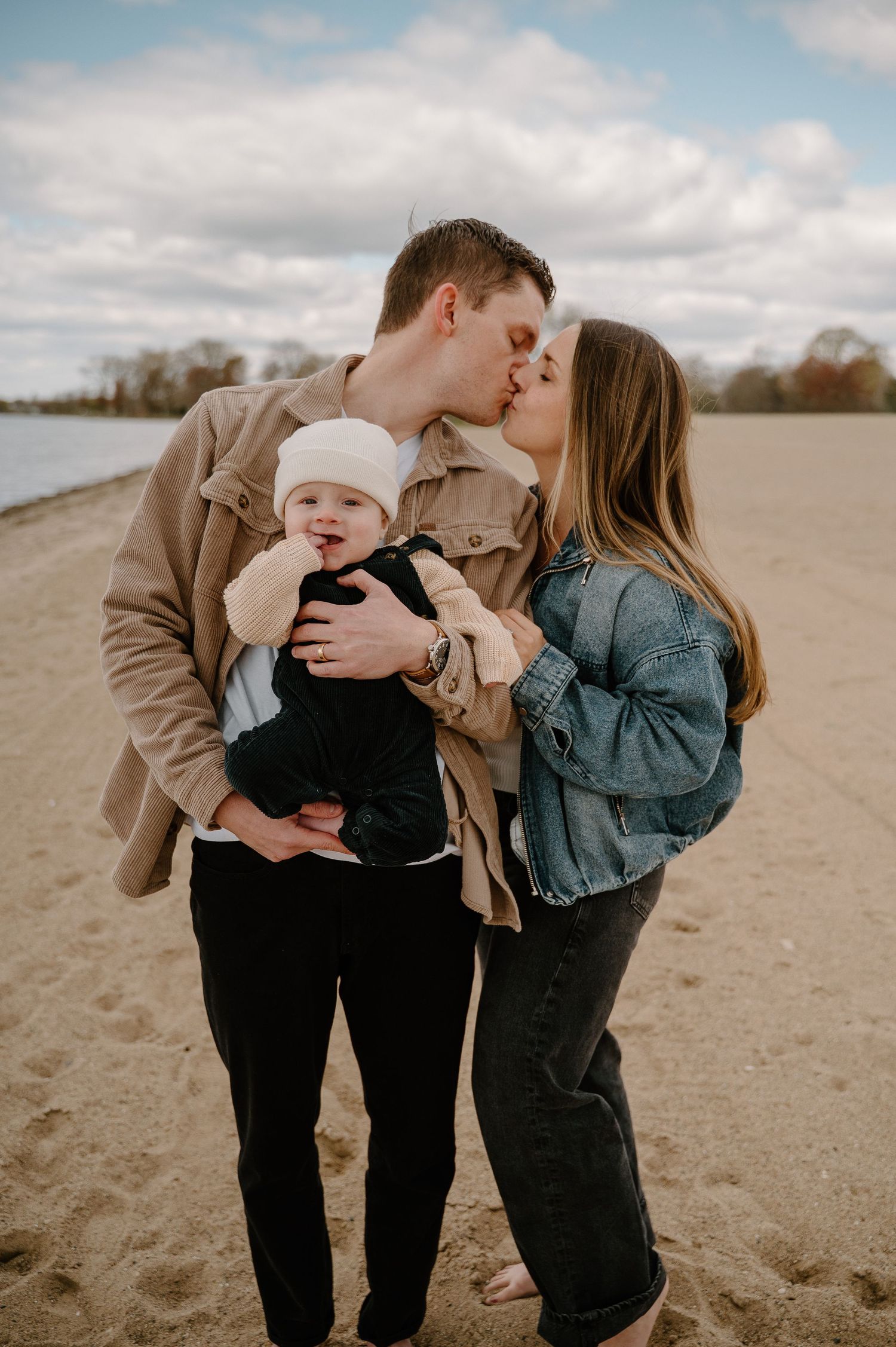 Young couple kissing on sandy beach while holding their smiling baby dressed in white hat and black outfit.