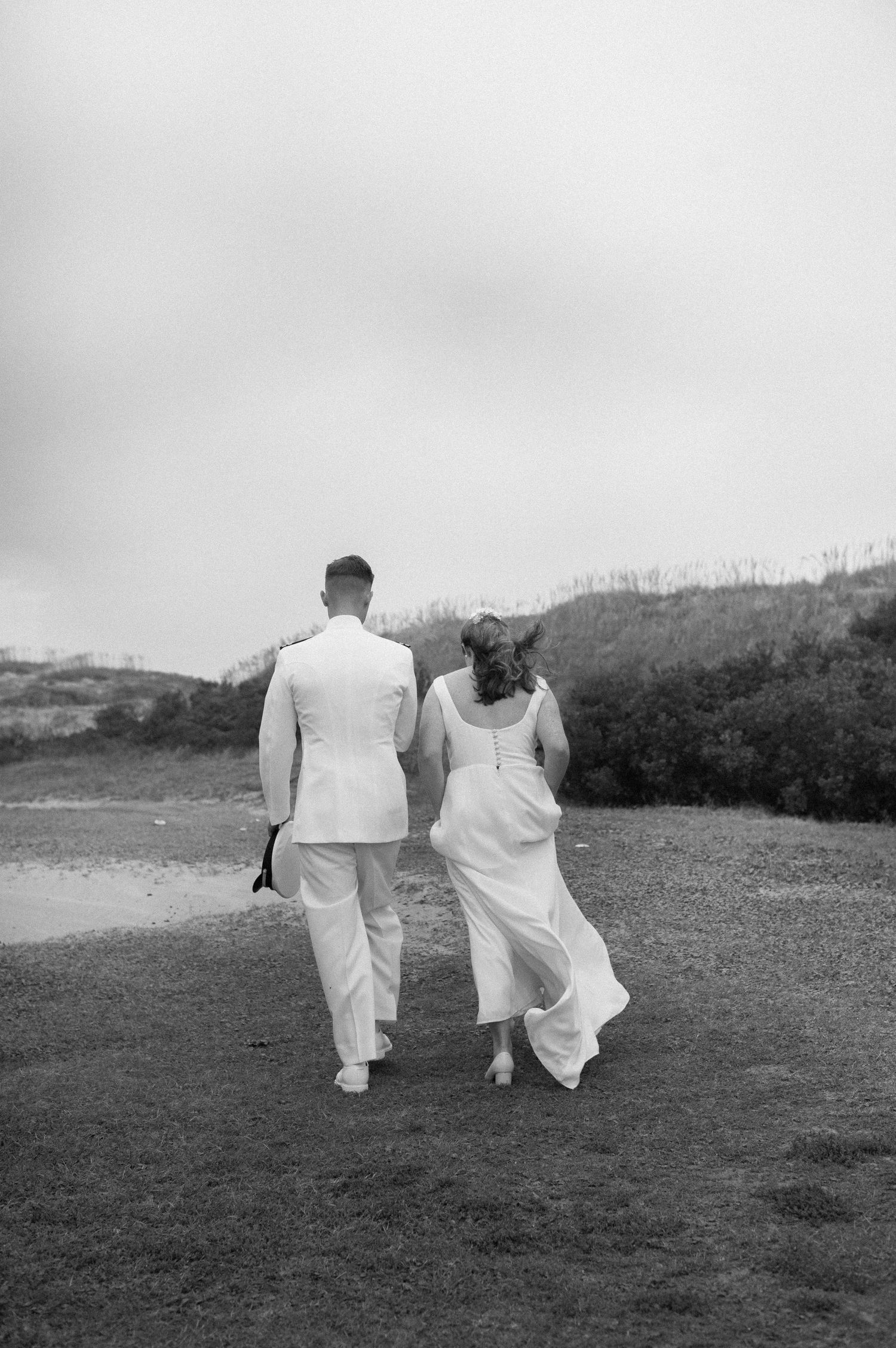 Black and white photo of a bride and groom walking away hand in hand across a grassy landscape.