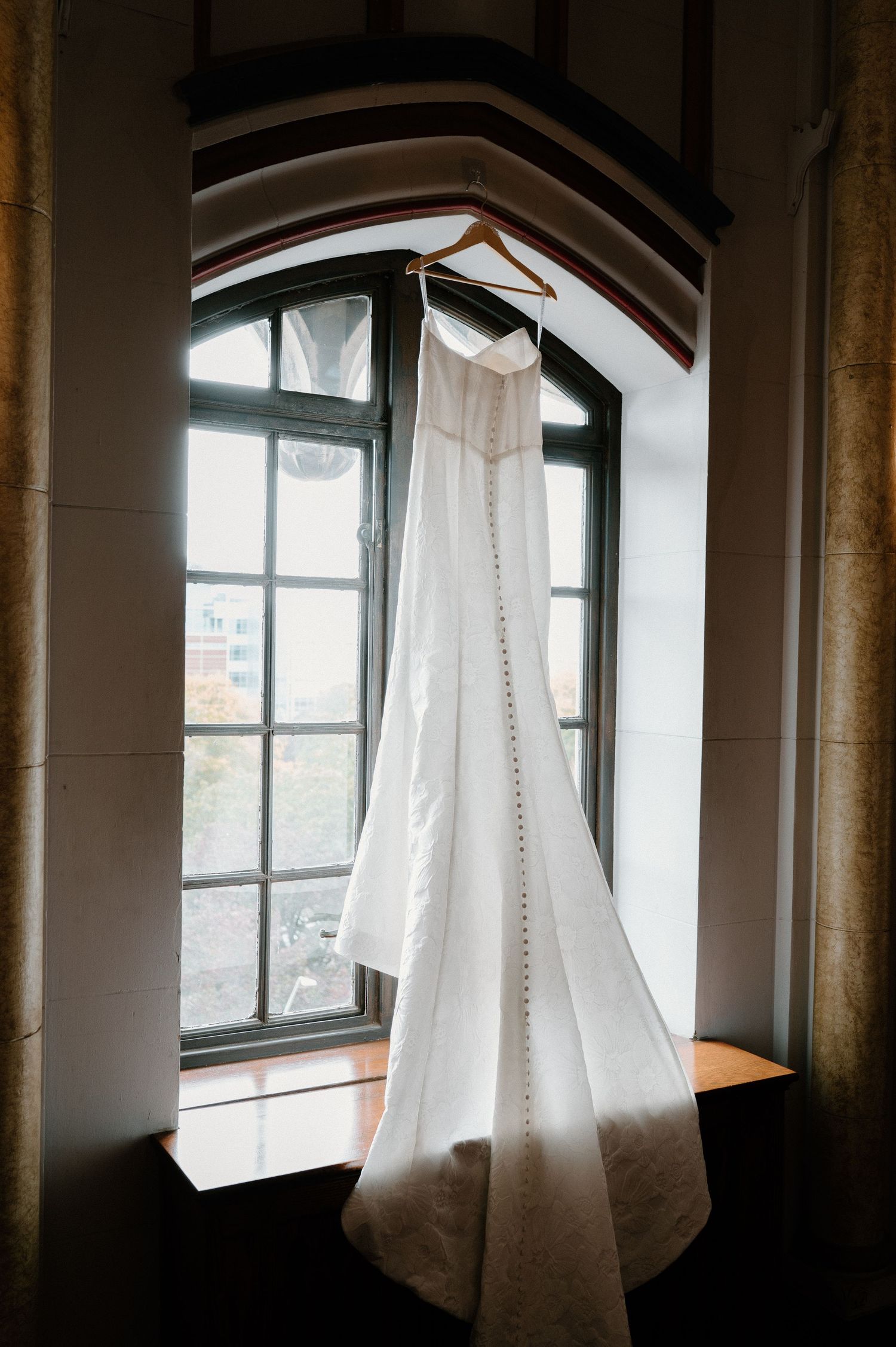 White wedding dress hanging on wooden hanger in arched window with button detail train.