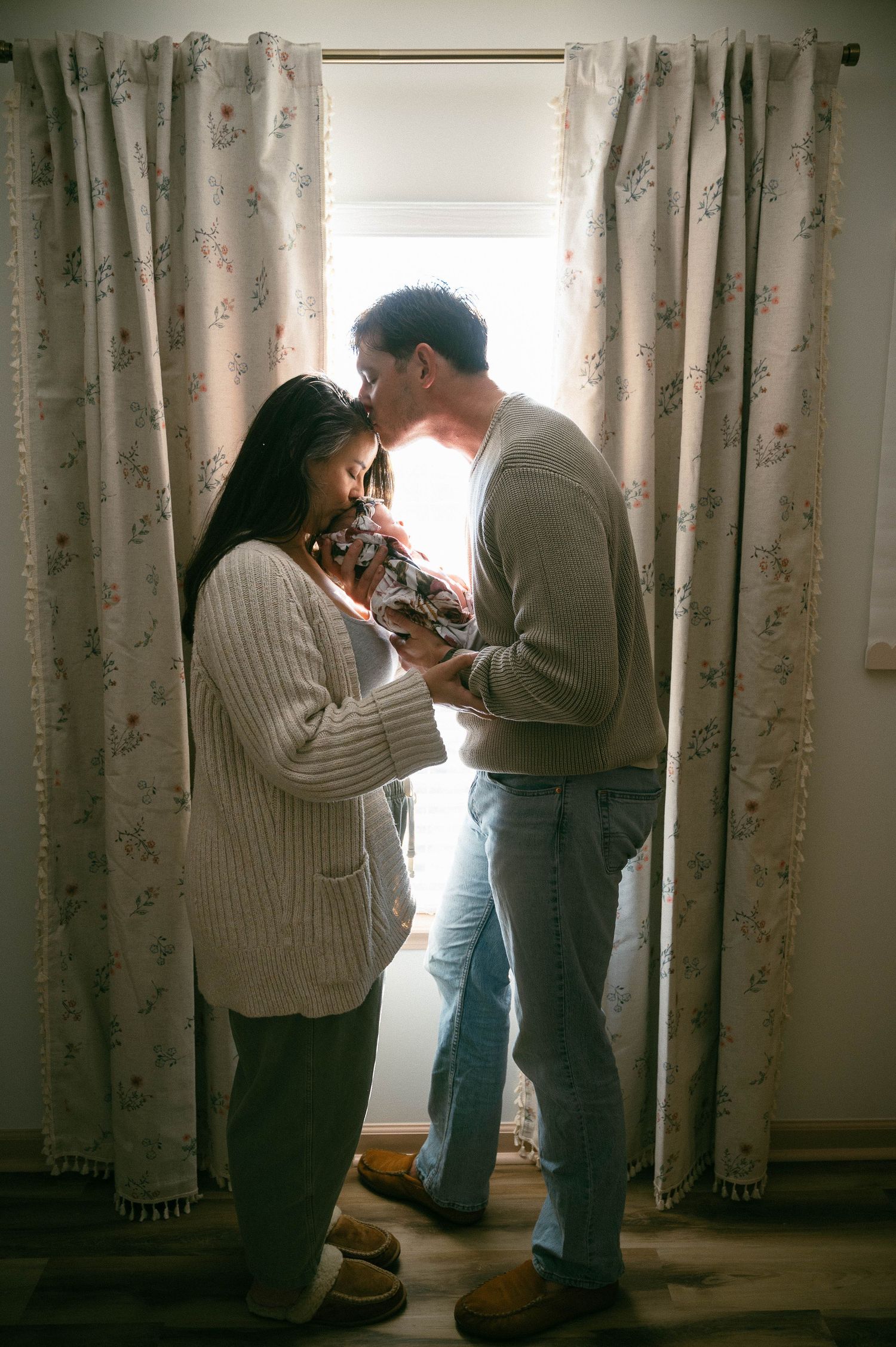 A couple stands by a sunlit window holding their newborn baby, sharing a tender moment in a cozy room.