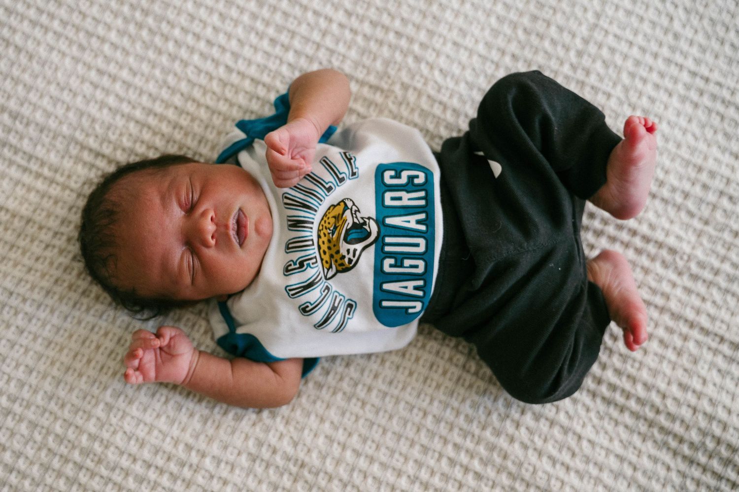 Newborn baby wearing a Jacksonville Jaguars bib and outfit lying on a white textured blanket.