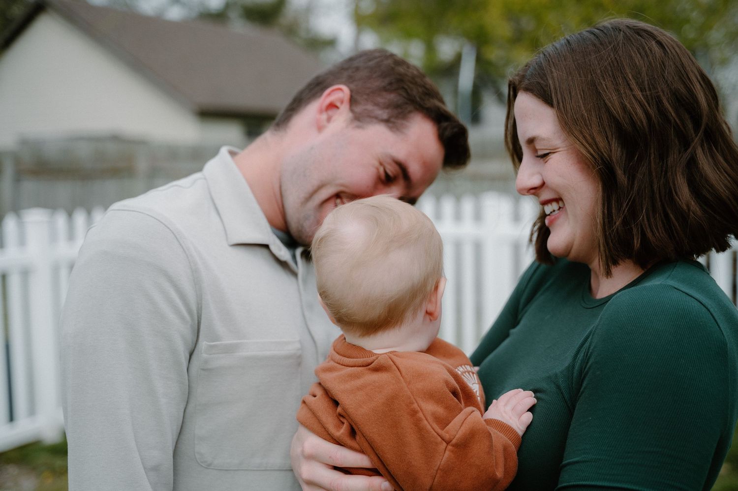 A smiling couple shares a tender moment with their baby outdoors near a white picket fence.