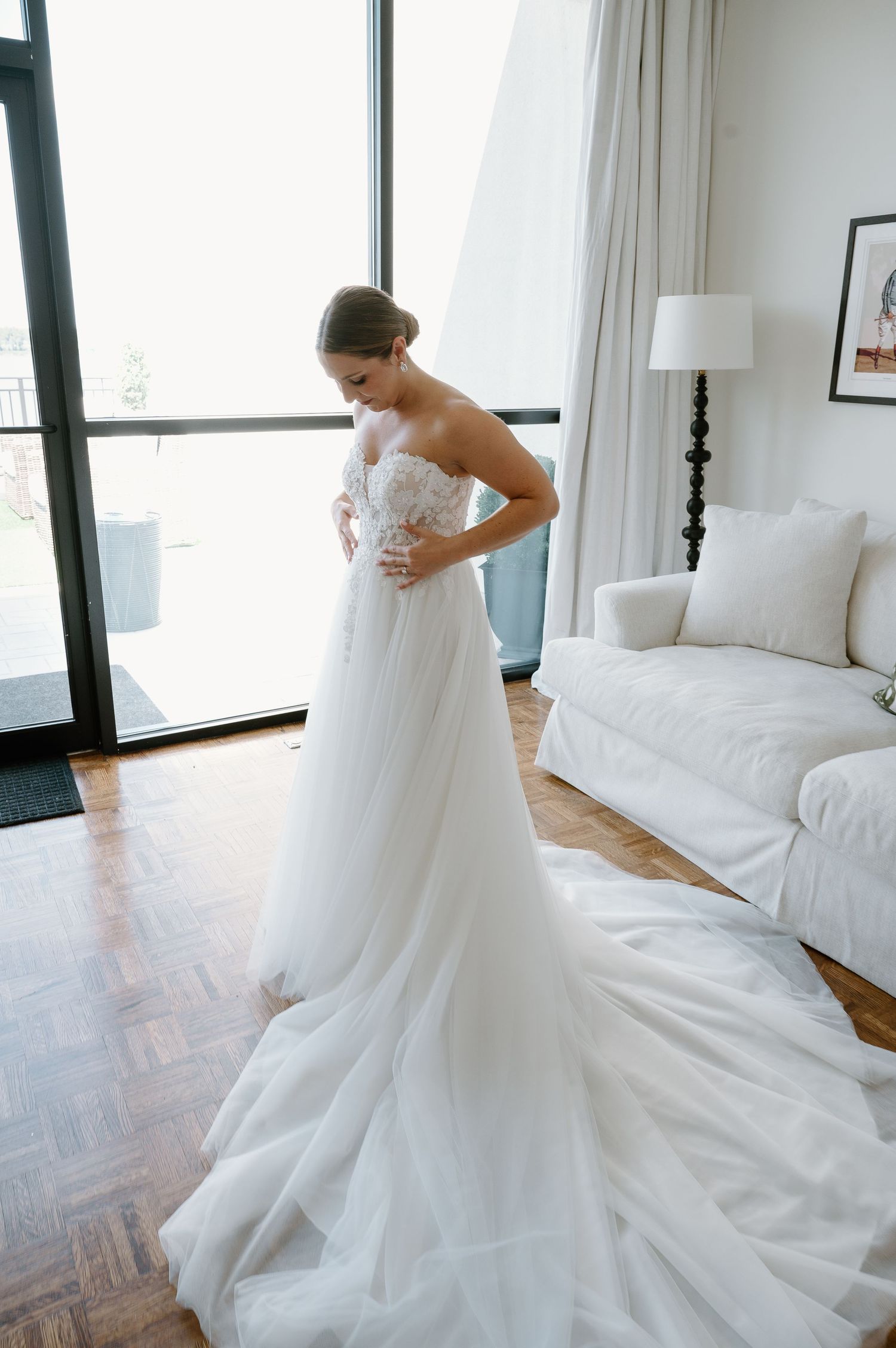 Bride in a strapless white tulle wedding gown with lace bodice stands in an elegant room with large windows.