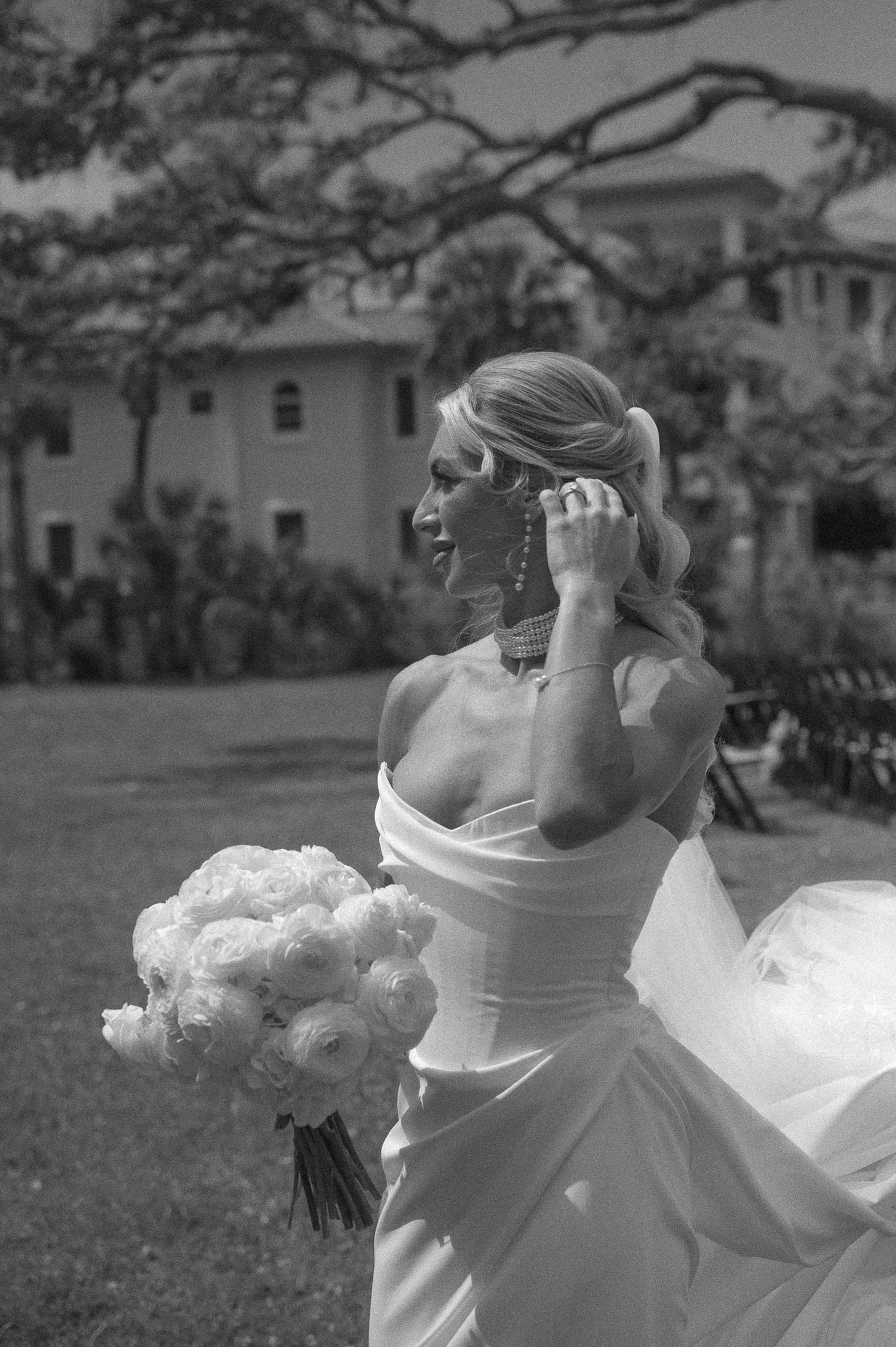 Bride in strapless white gown holding peony bouquet, touching earring outdoors, black and white wedding photo.