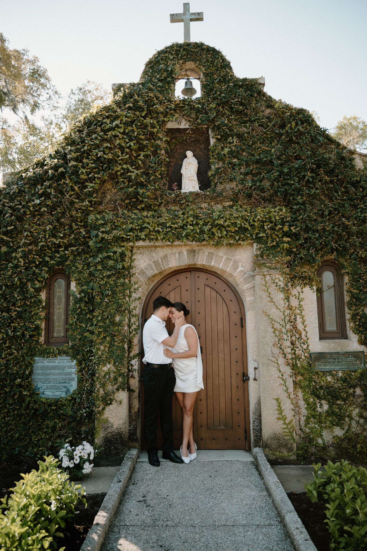 Couple sharing romantic moment in front of ivy-covered chapel with cross and wooden arched door.