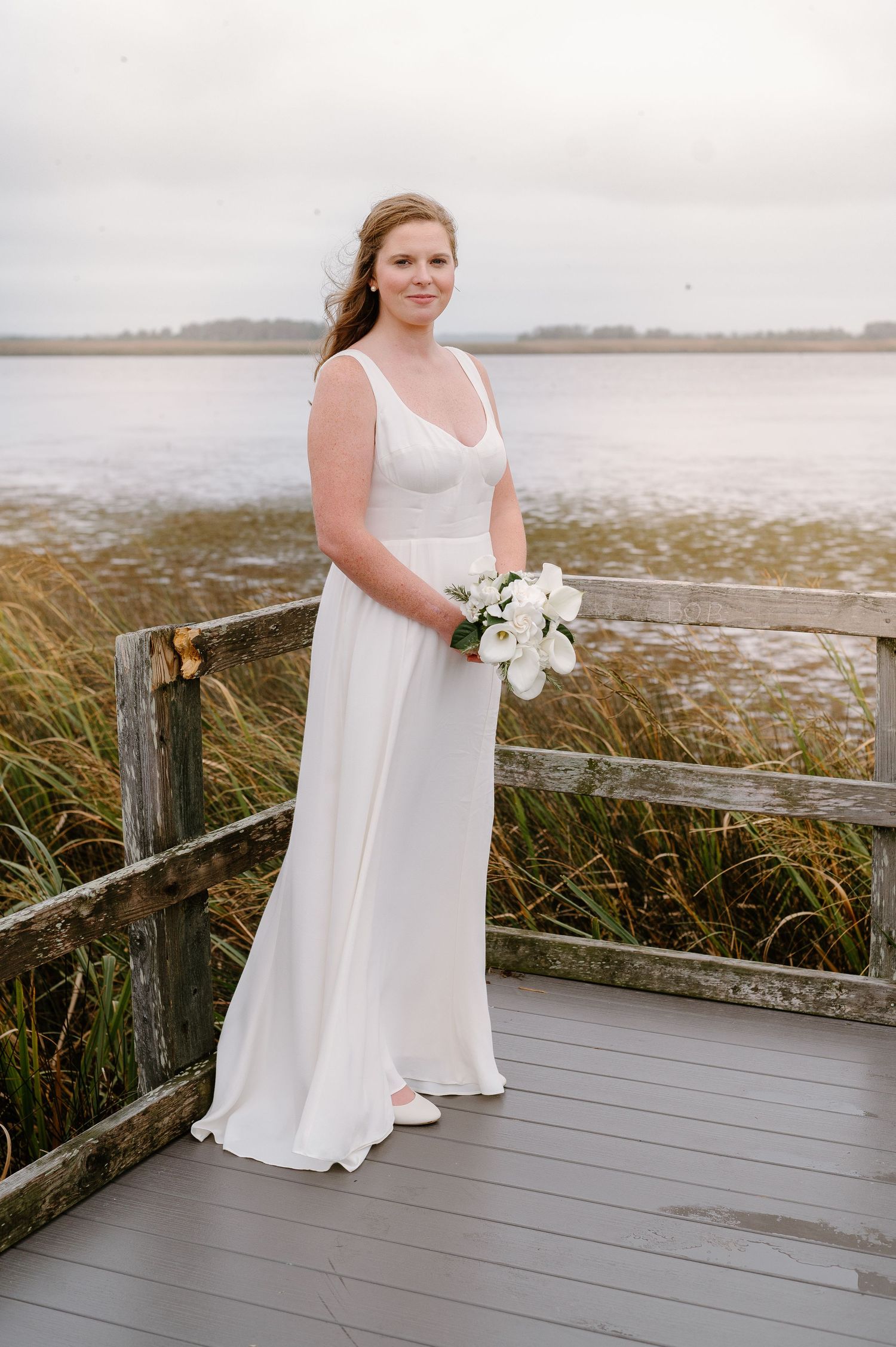 Bride in elegant white dress poses on wooden boardwalk near coastal marshland.