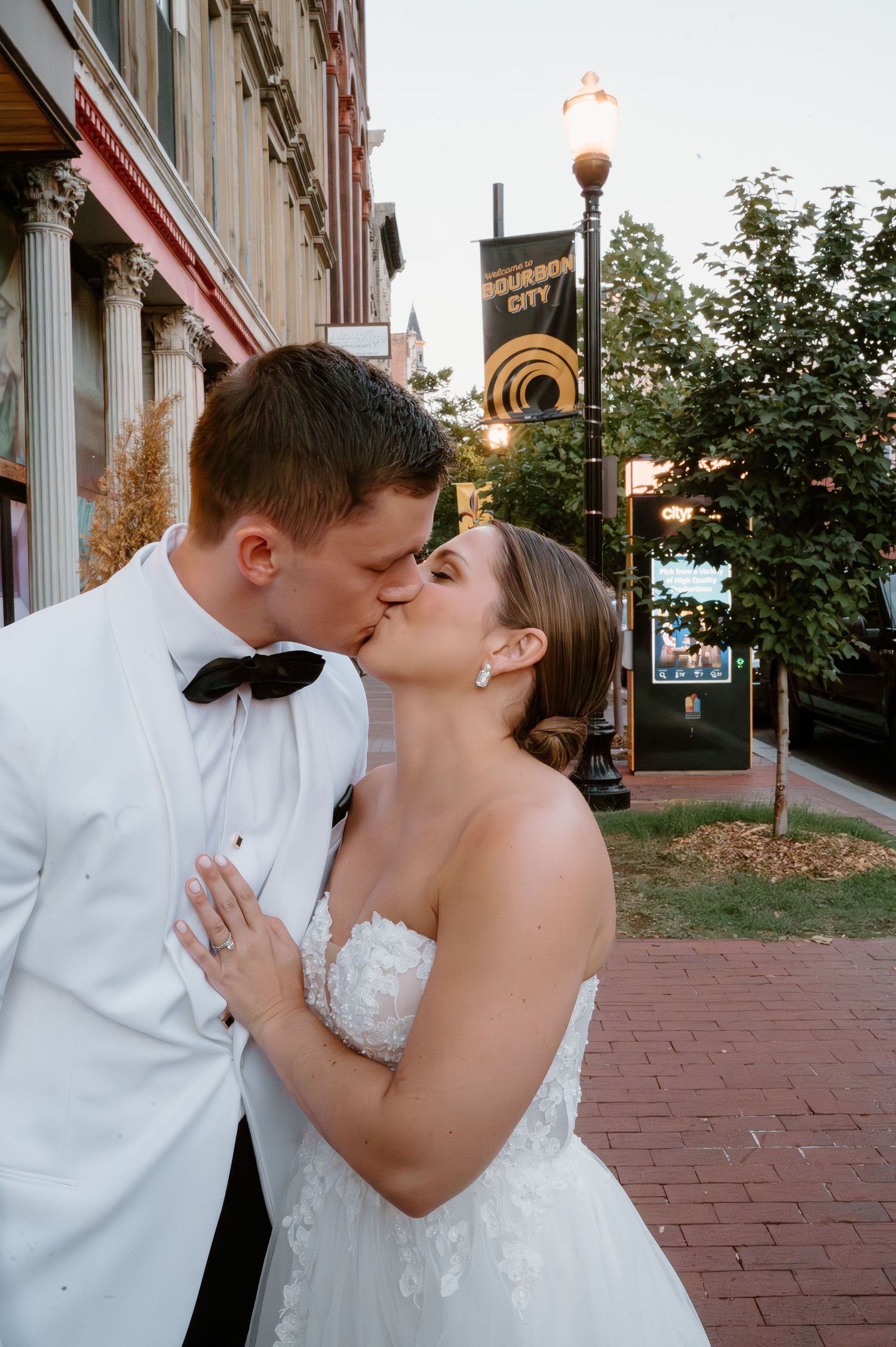 Romantic wedding scene on a brick walkway with decorative streetlights and architecture in the background.