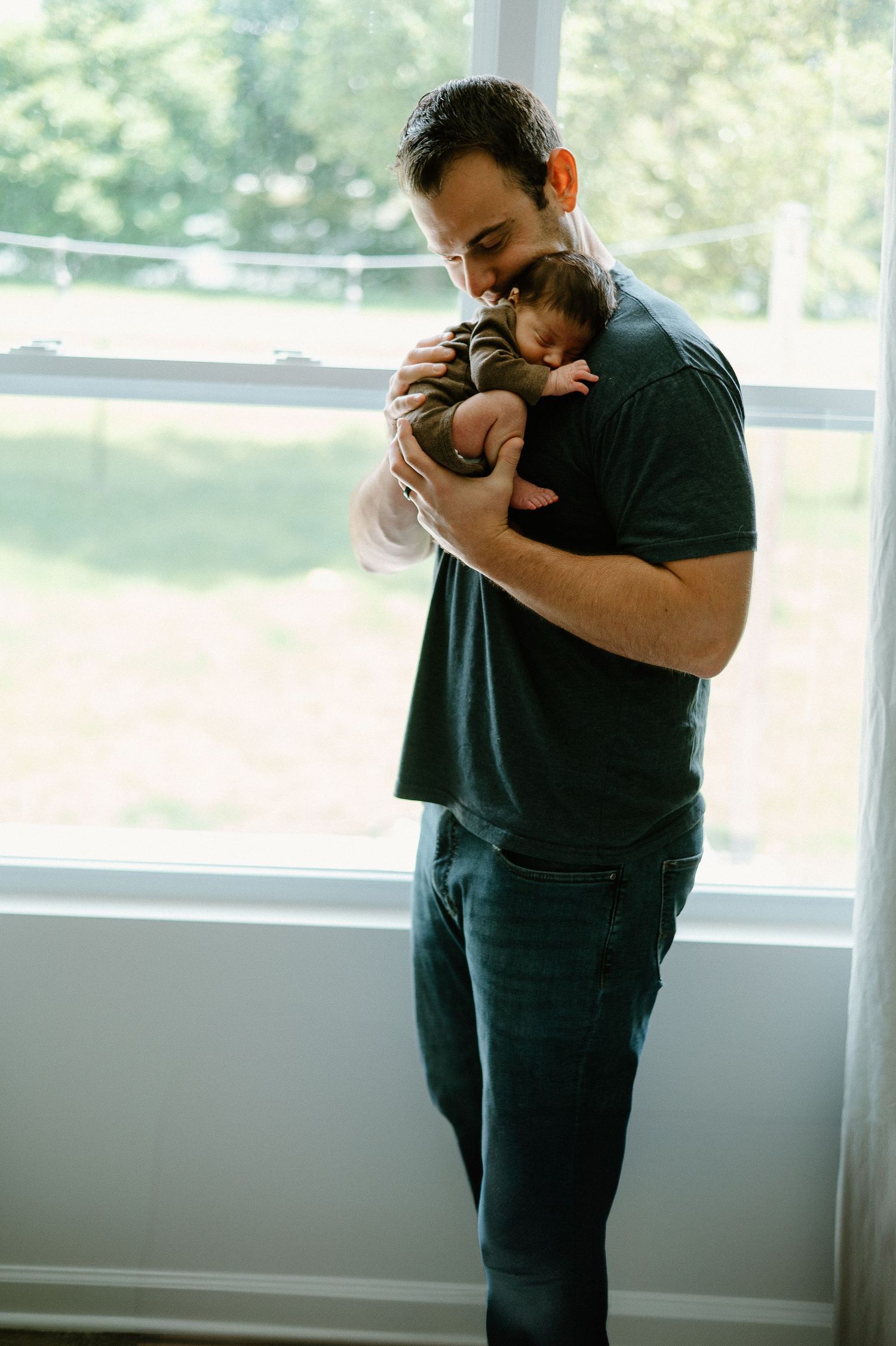 A parent holds a sleeping infant near a bright window.