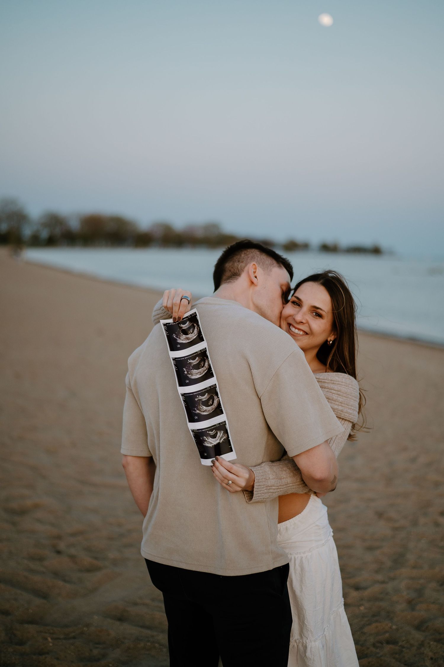 Romantic beachside pregnancy announcement photos featuring ultrasound prints at dusk.