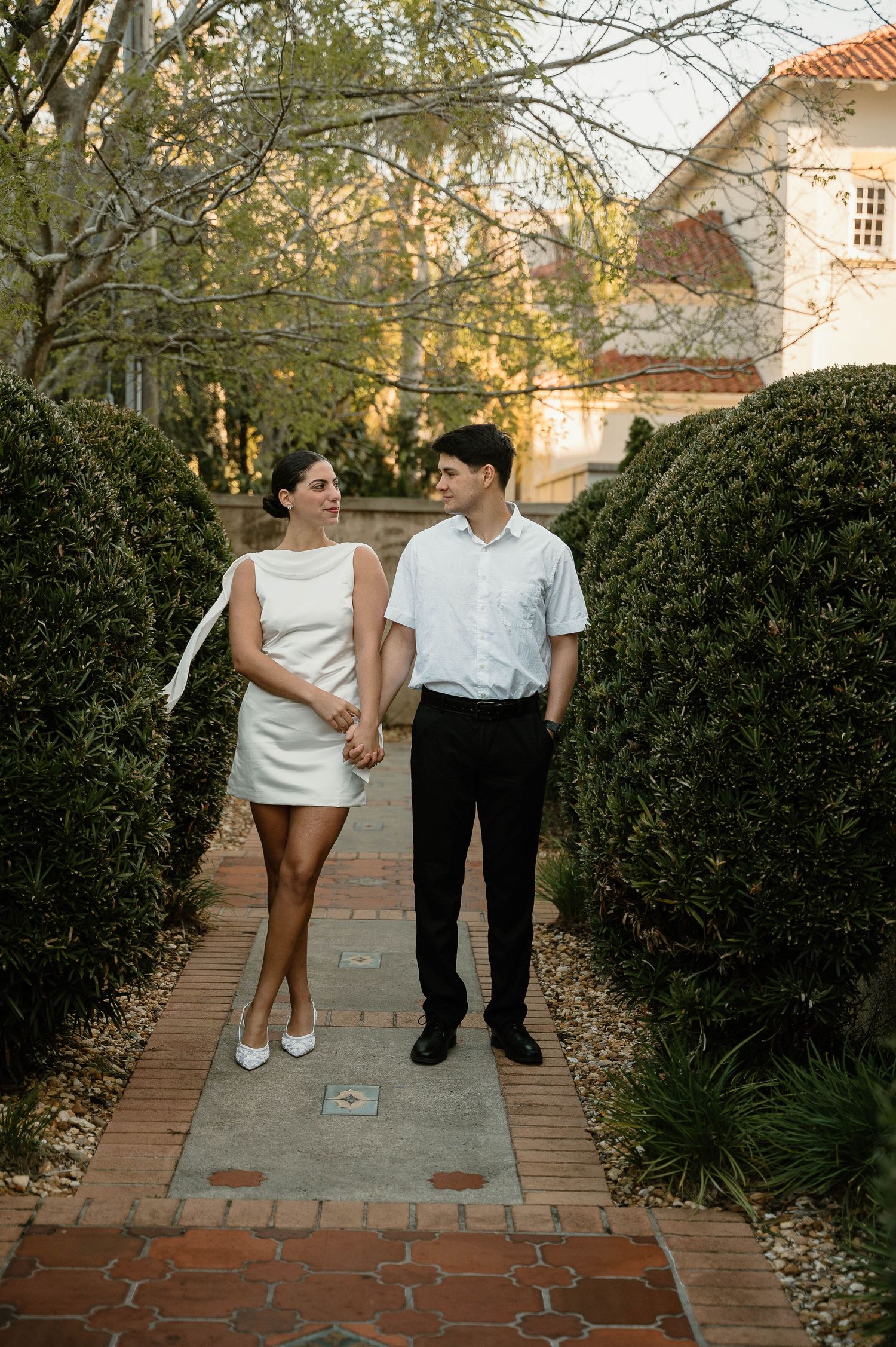 Couple holding hands walking on a brick garden path, woman in white mini dress, man in black pants and white shirt.