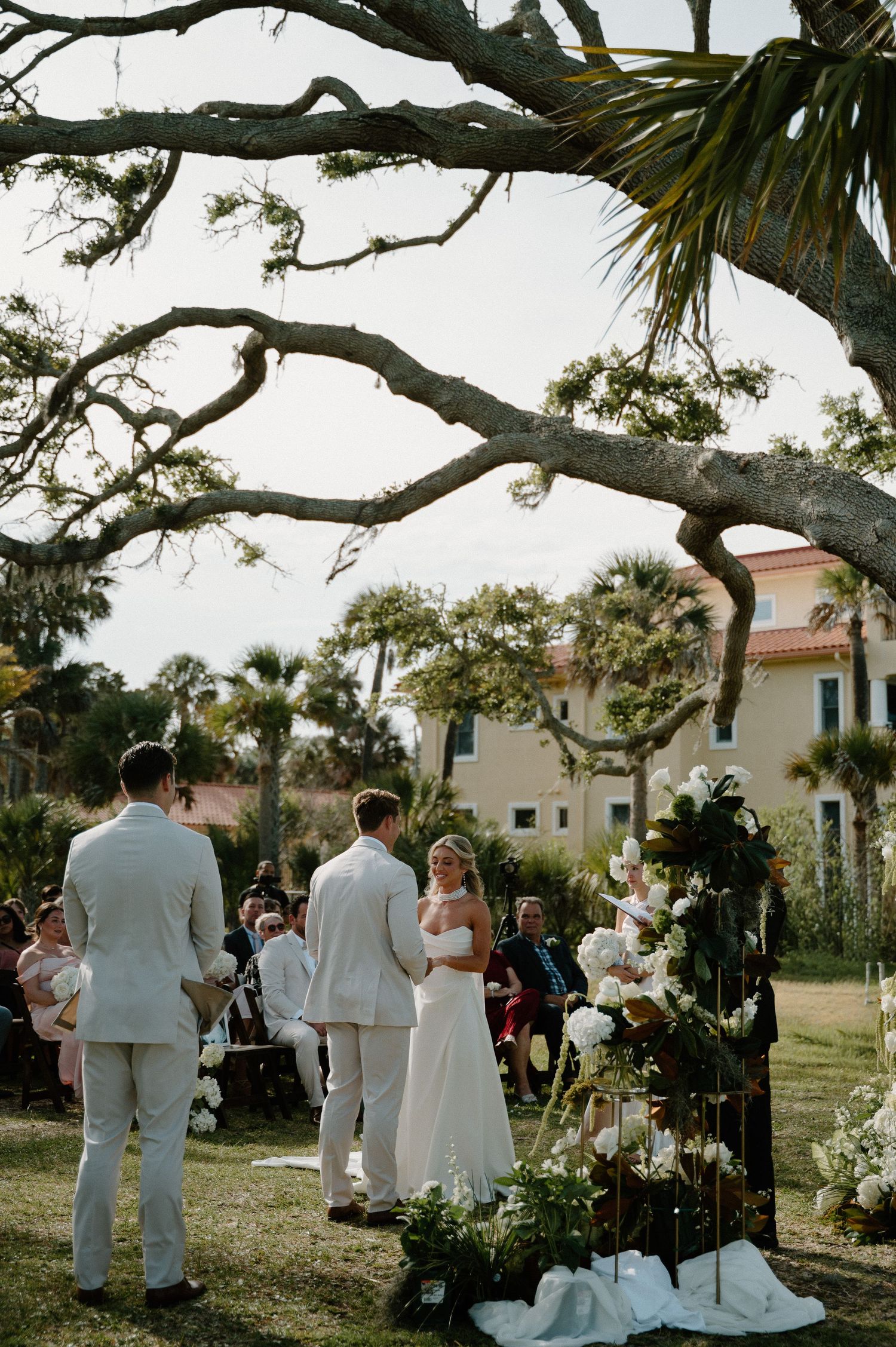 Outdoor wedding ceremony under sprawling oak tree with couple at floral arch, guests seated on lawn near Mediterranean-style building.