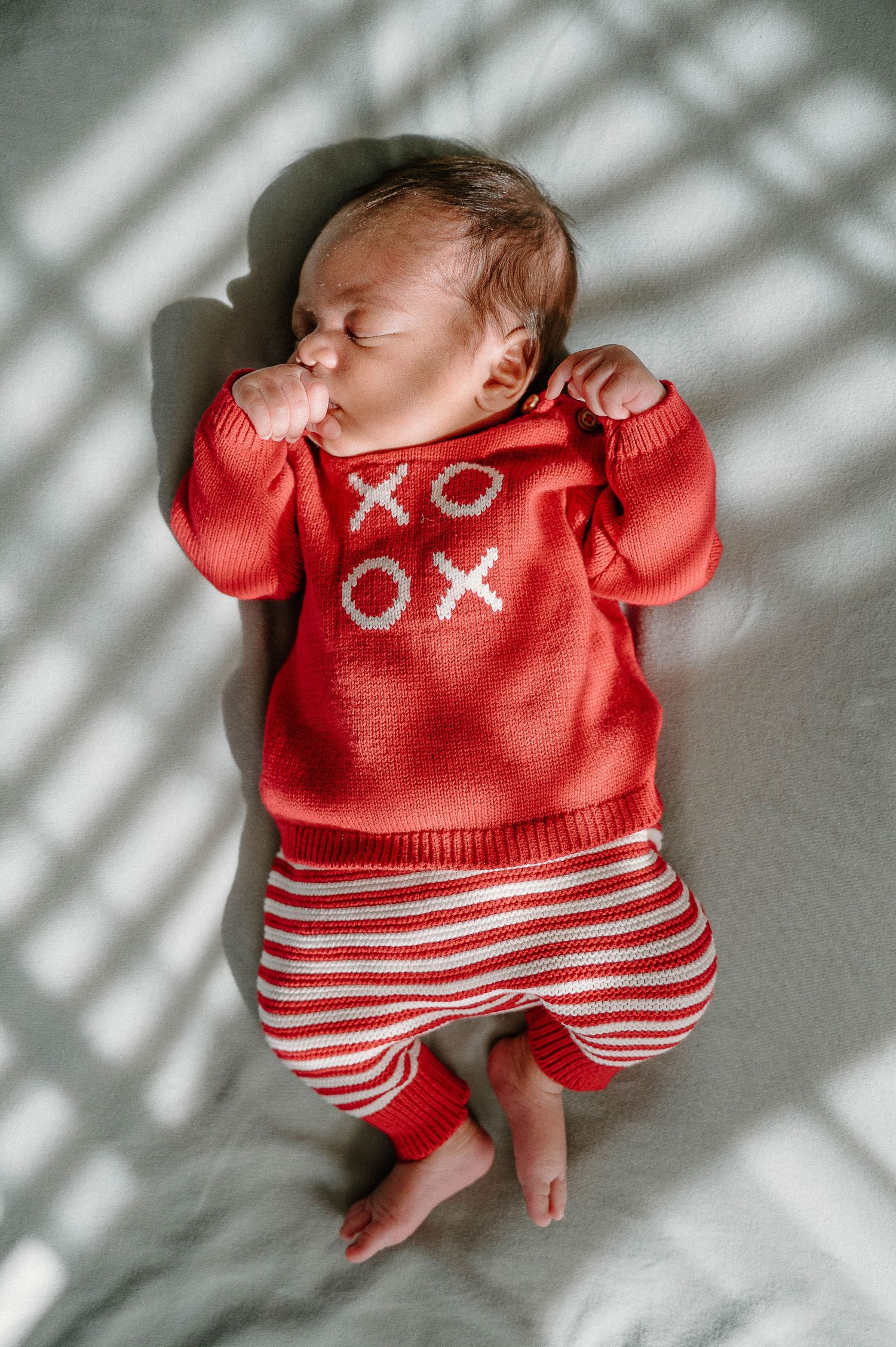 Sleeping newborn baby wearing a red XOXO knit sweater and red striped pants lying on a white blanket.