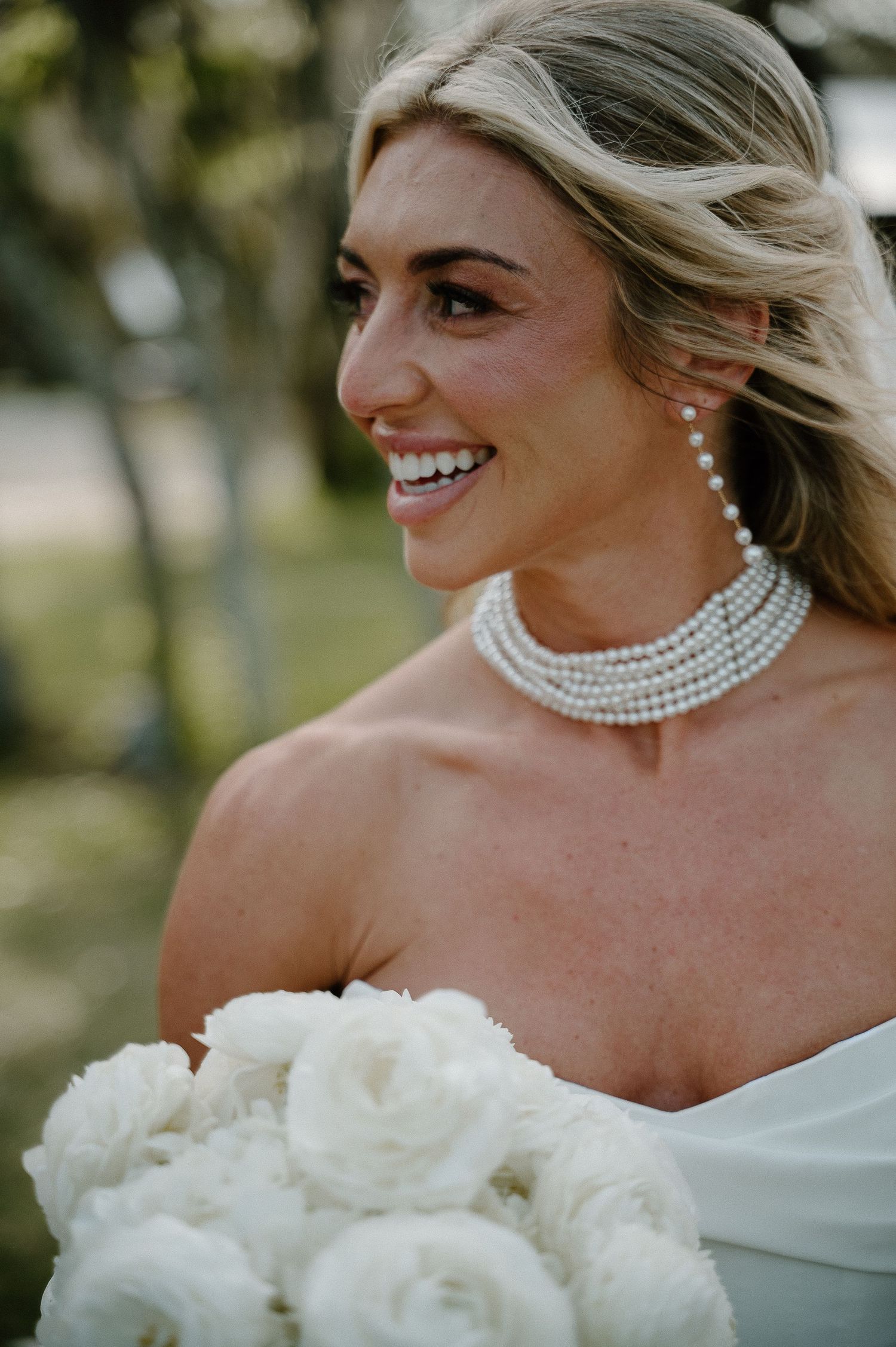 Smiling bride in strapless white gown with pearl choker and earrings holding white rose bouquet outdoors.