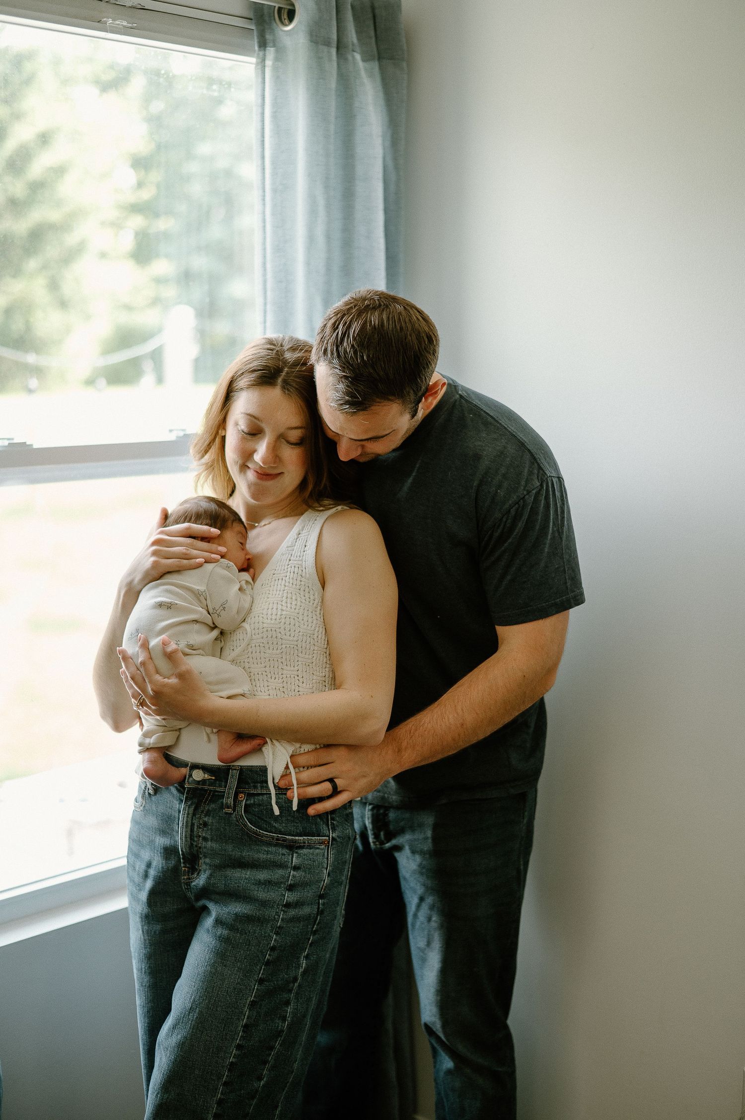 A couple shares an intimate moment with their newborn by a bright window.