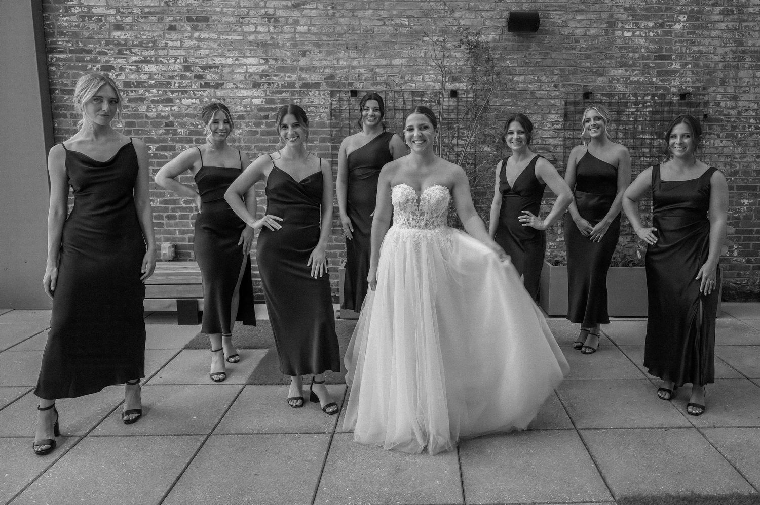 Wedding party lineup in black dresses against an industrial brick wall.
