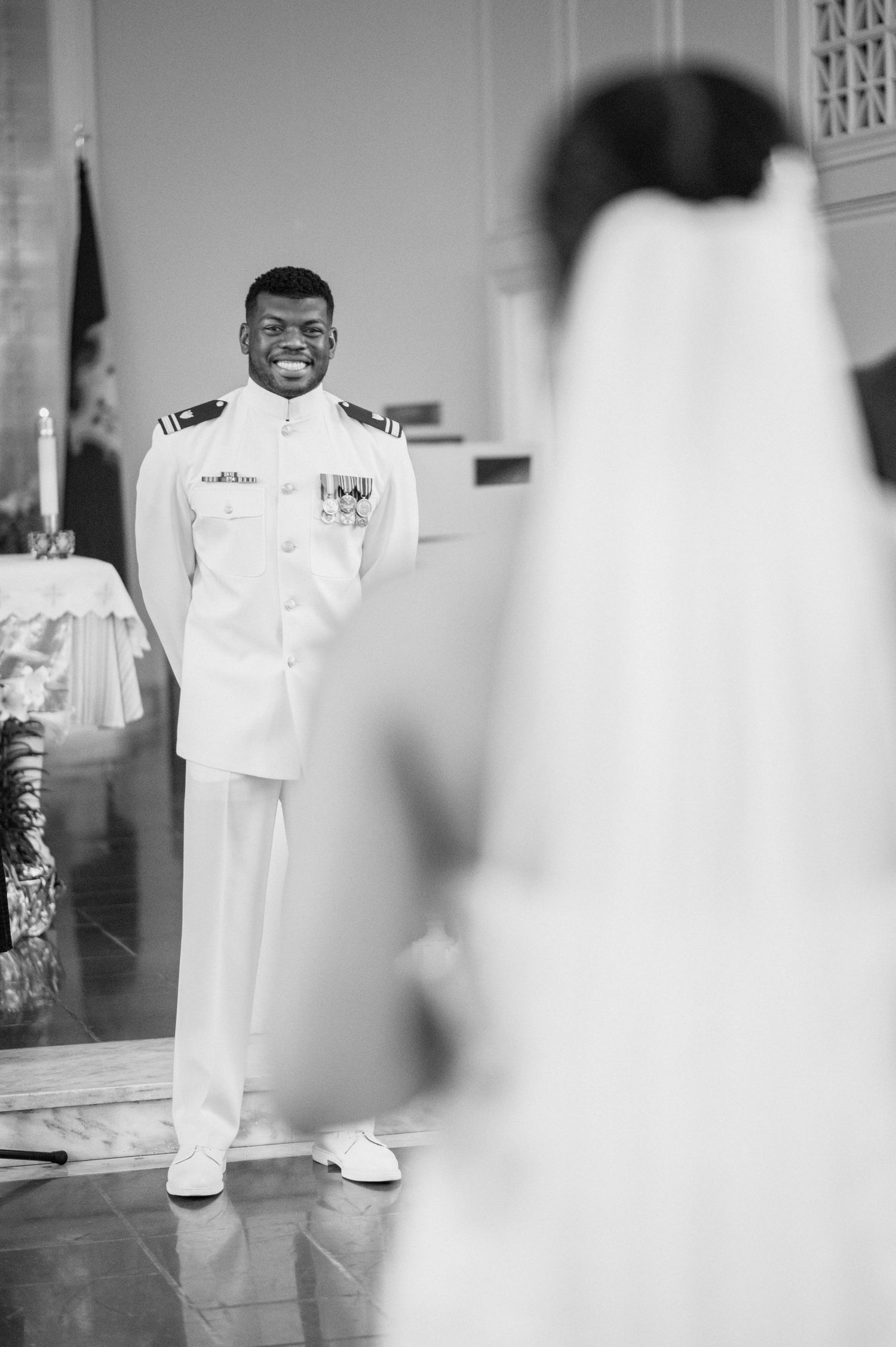A joyful figure smiling in a crisp white formal uniform stands in a hallway.