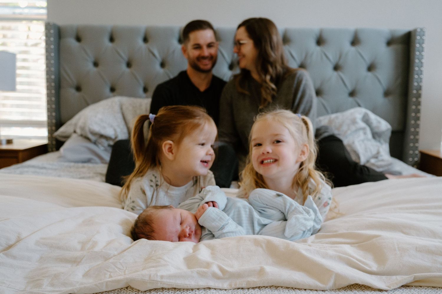Family of five with two smiling girls holding newborn baby on bed, parents sitting behind them smiling.