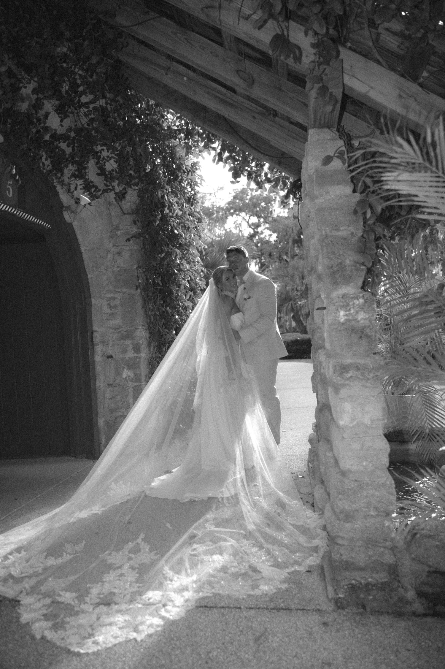 Black and white wedding photo of bride in long veil and groom embracing under stone arch surrounded by lush greenery.