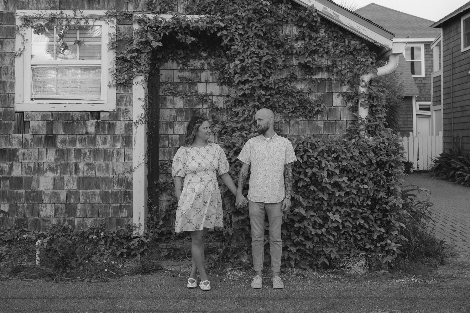 Black and white photo of a couple holding hands in front of an ivy-covered house.